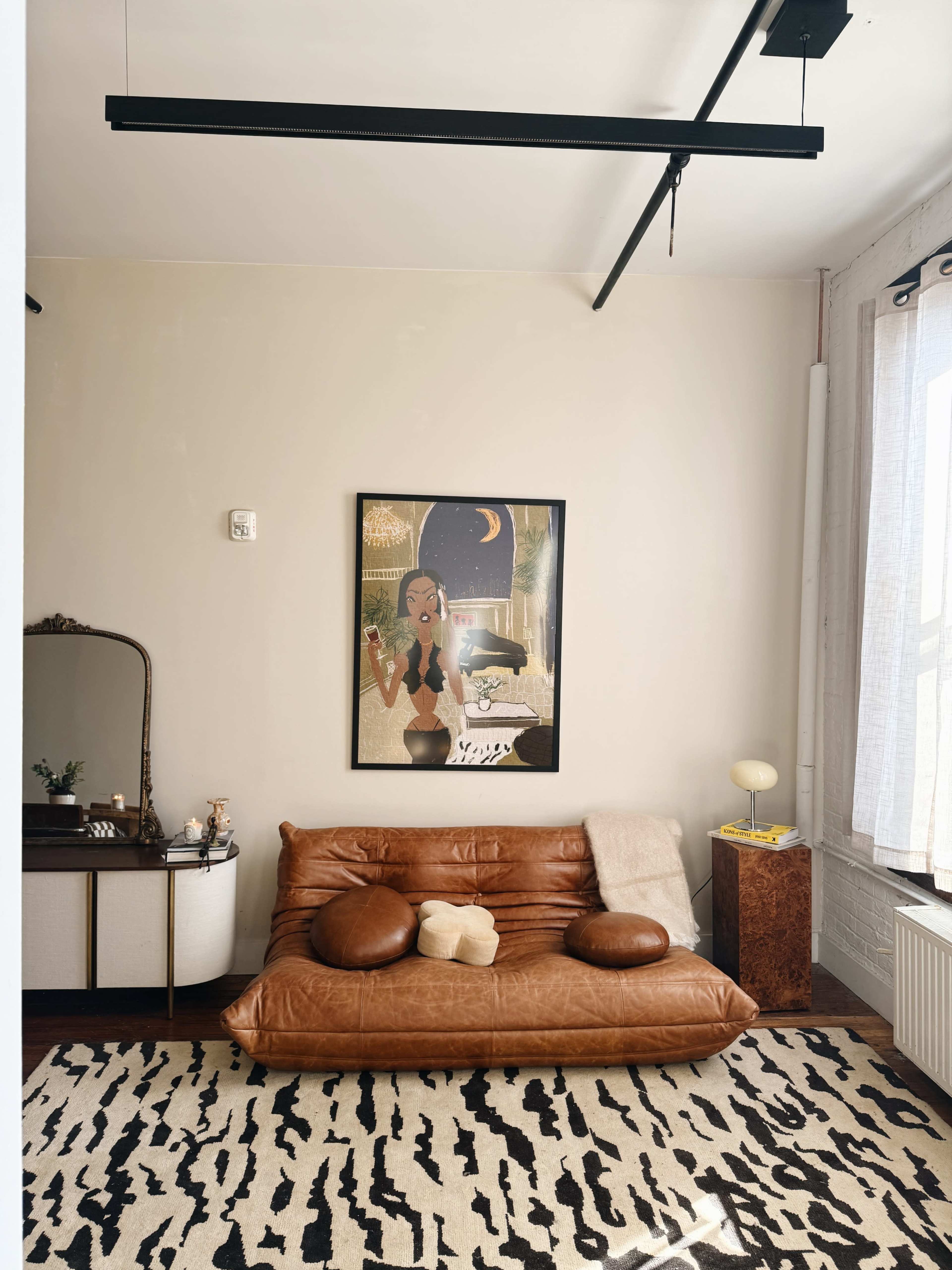 A cozy room features a brown leather sofa, a vintage dresser, and a large framed artwork on the wall.