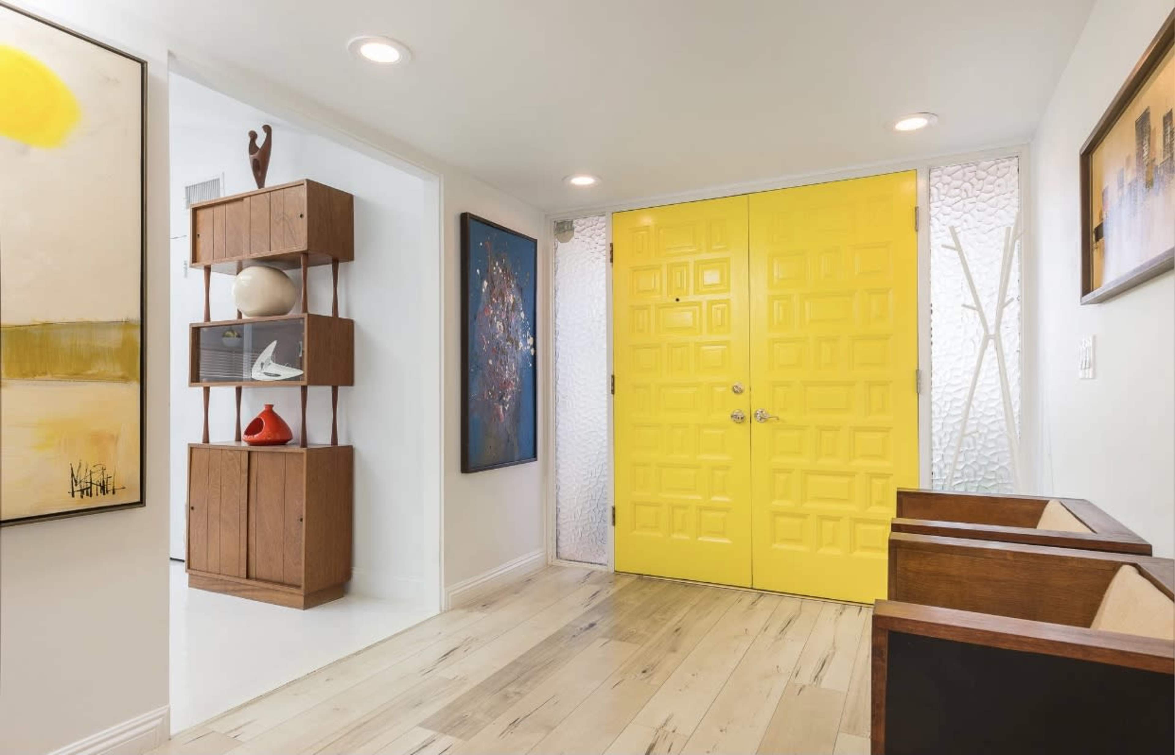 The image shows a modern entryway featuring yellow double doors, wooden furniture, and abstract artwork on the walls.