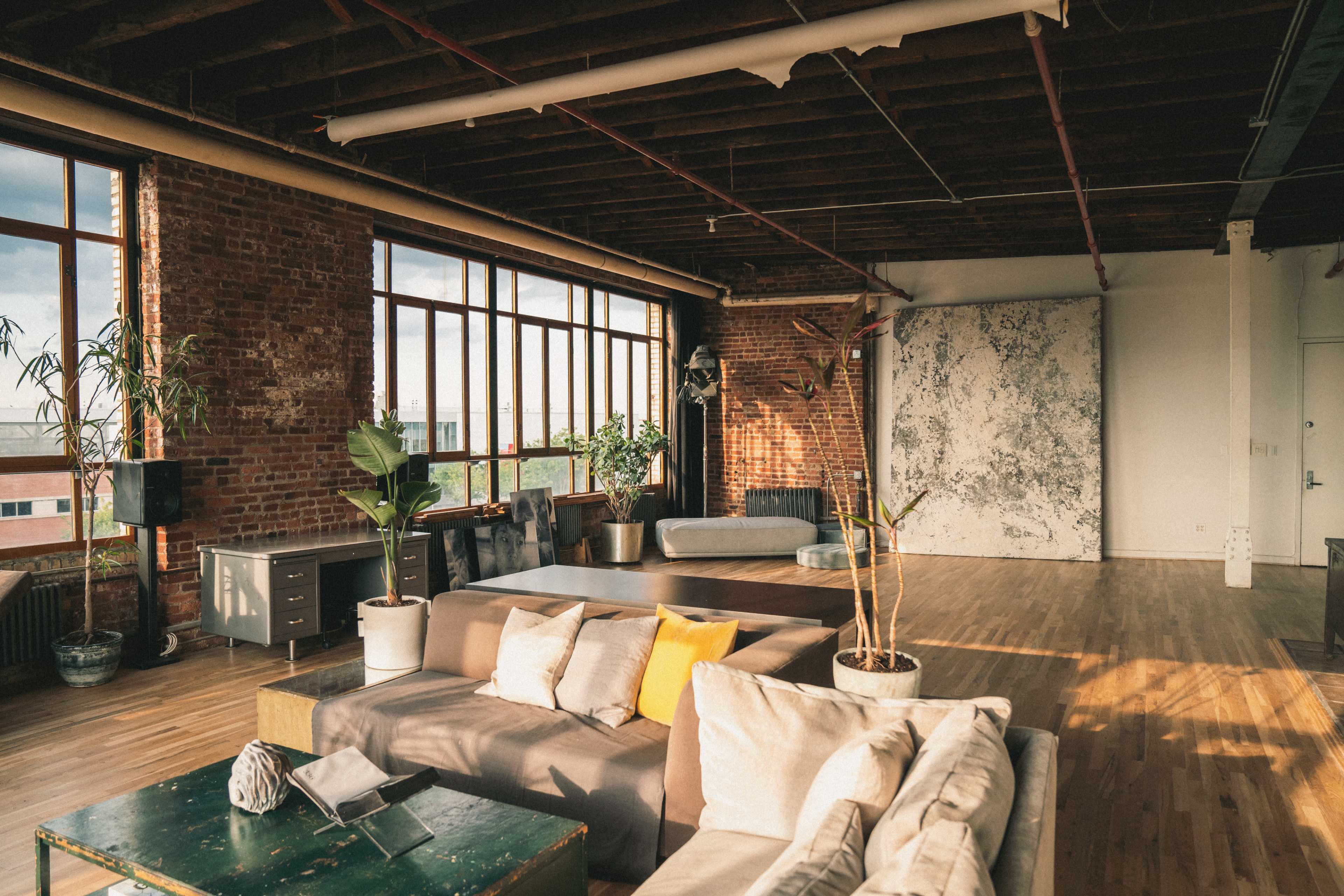 The image shows a spacious, industrial-style living area with large windows, brick walls, and multiple seating arrangements.