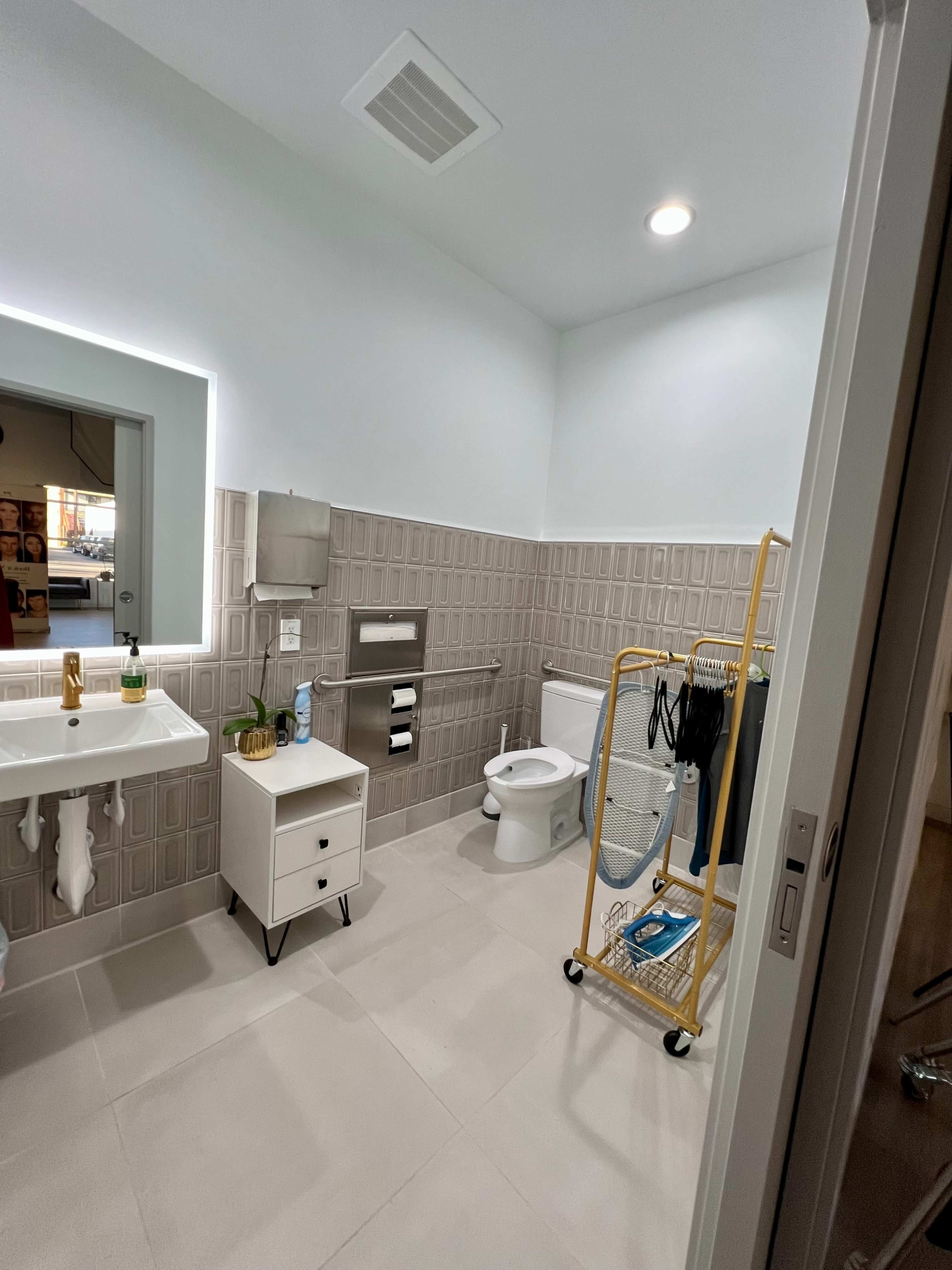 A modern bathroom featuring a sink, toilet, wall-mounted fixtures, and a clothing rack displaying garments.