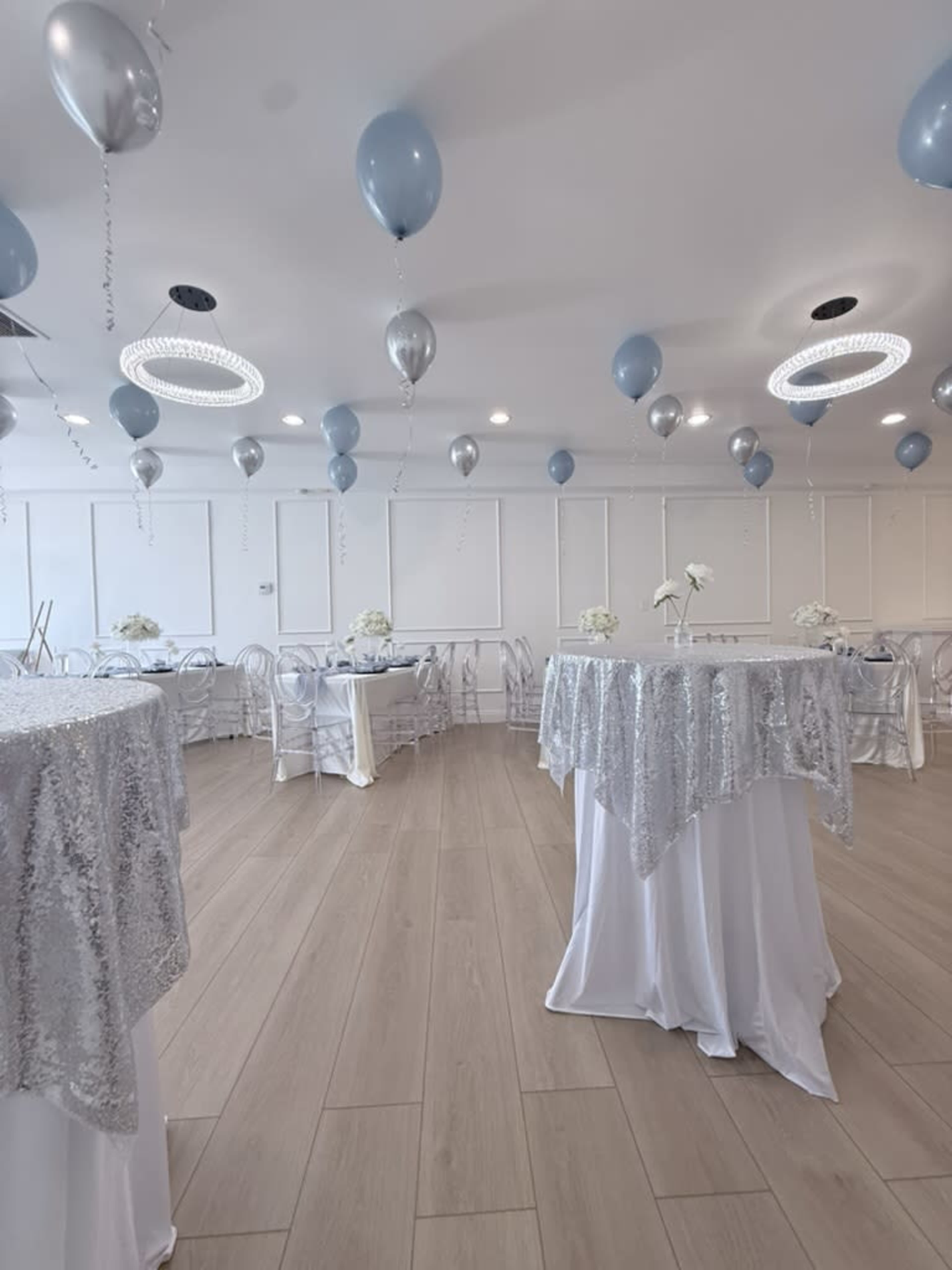 The image shows a decorated event space with round tables covered in silver sequin cloths, white chairs, and balloons hanging from the ceiling.
