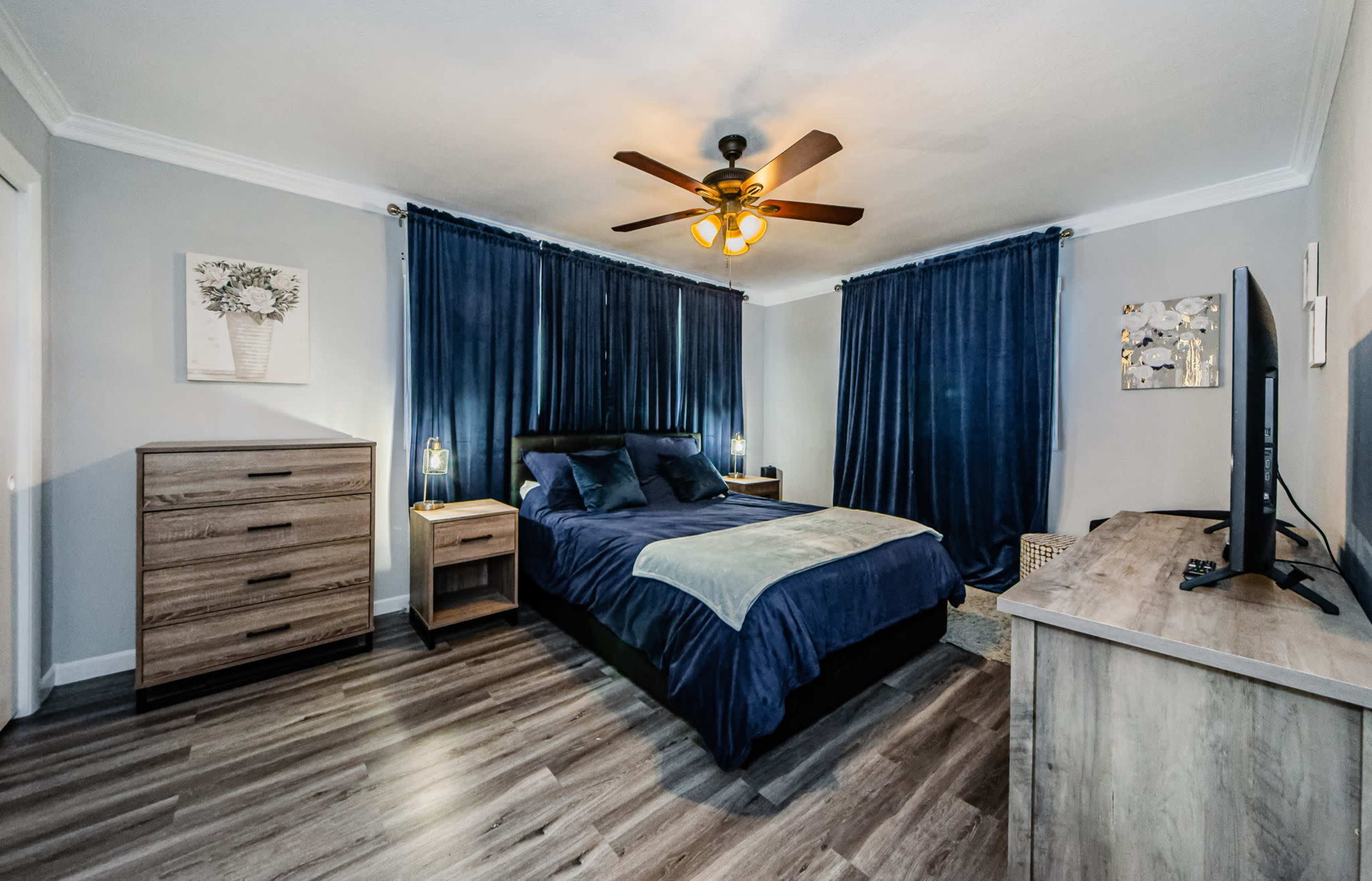 A modern bedroom features a large bed with dark bedding, wooden furniture, and blue curtains, illuminated by a ceiling fan with a light fixture.