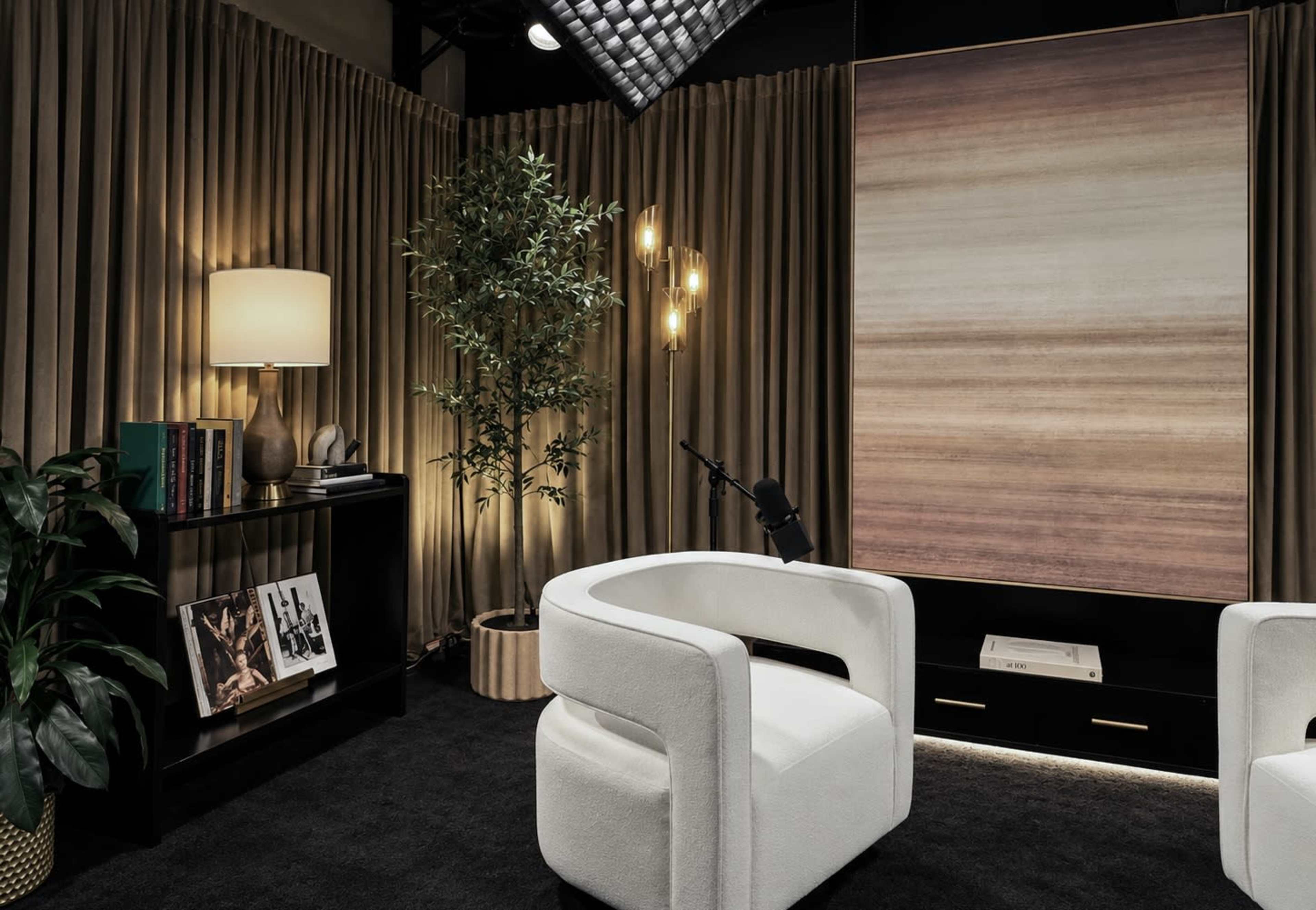The image shows a cozy interior scene featuring two white armchairs, a small black bookshelf with books and framed photos, a potted plant, and a decorative wall art piece, all set against a backdrop of golden curtains.