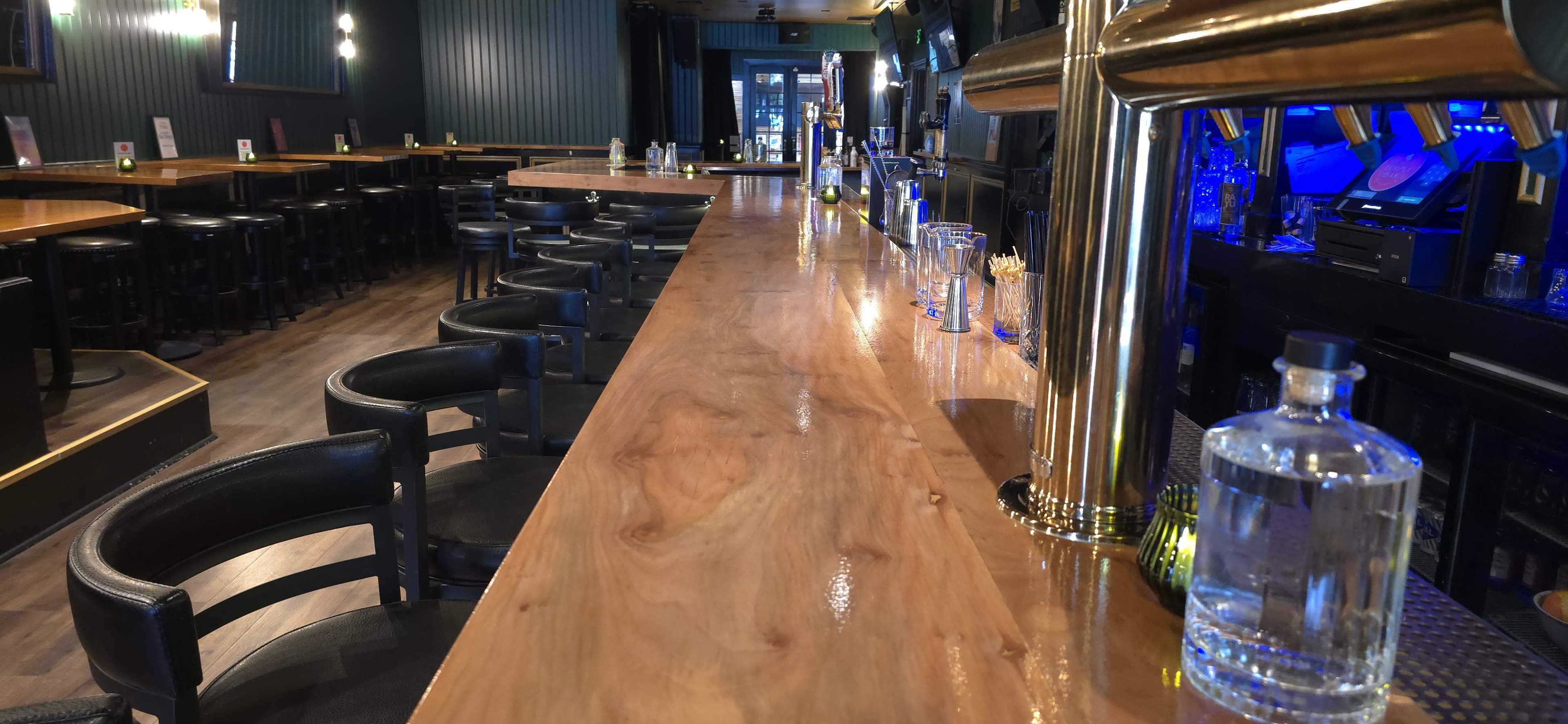 The image shows a well-lit bar area with a polished wooden counter, black leather stools, and a variety of glassware and bottles lined up along the back.