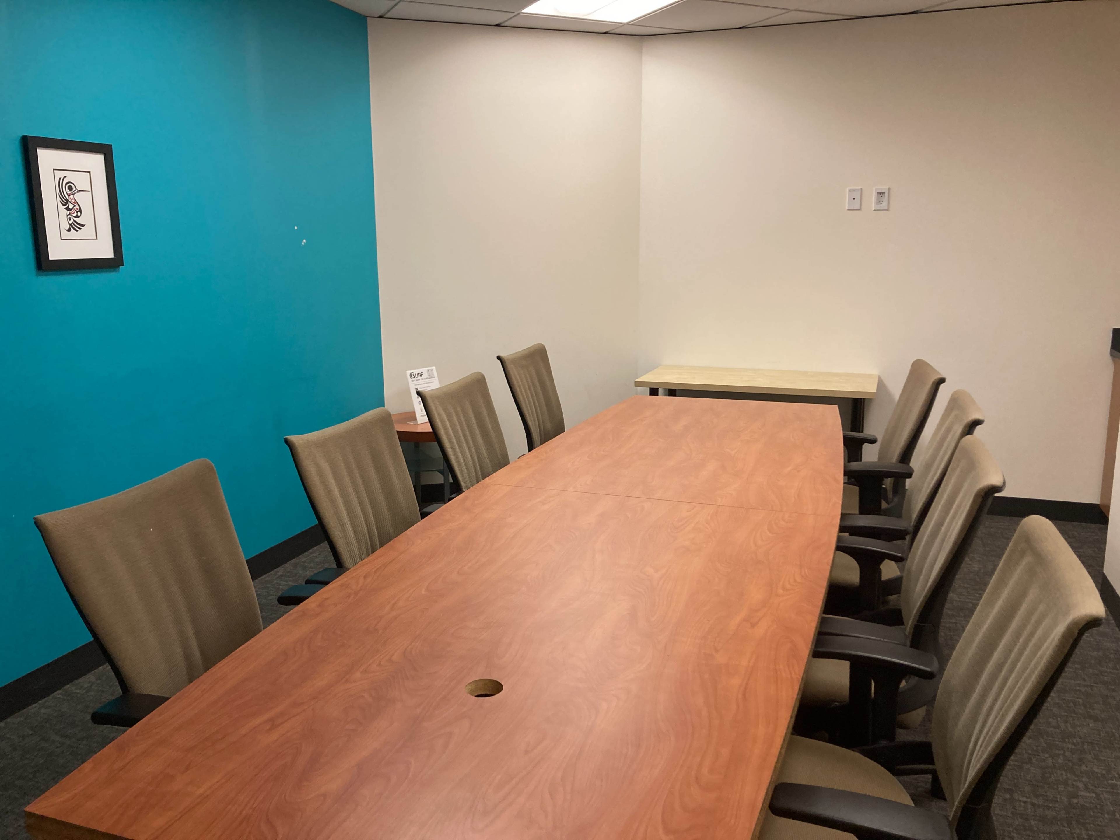 A conference room features a long wooden table surrounded by six chairs, with a blue accent wall and a small table in the corner.