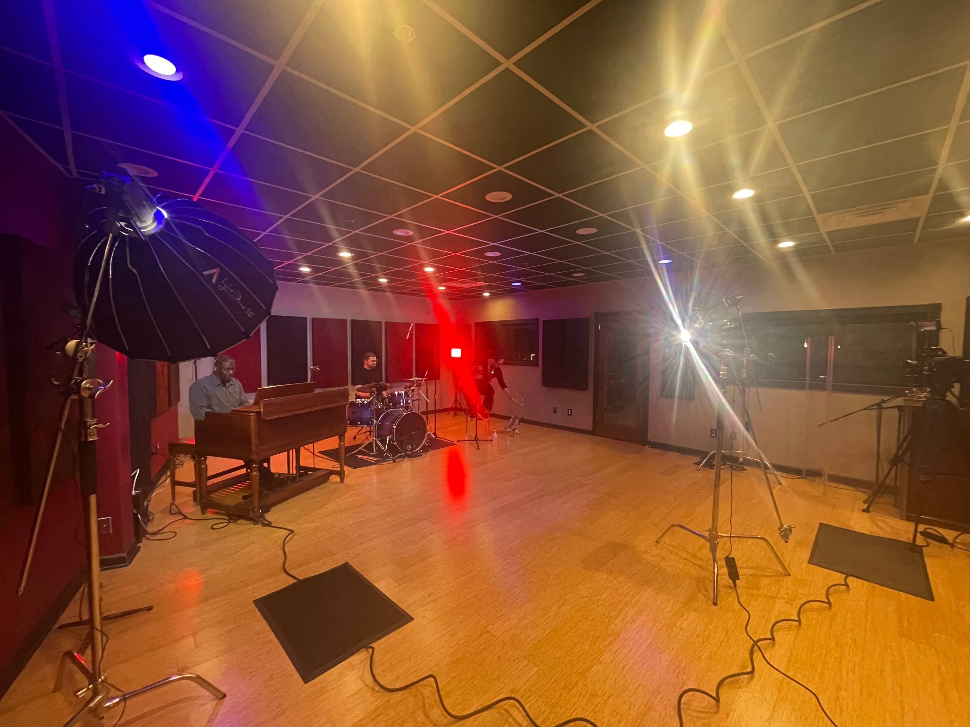 A music recording studio is set up with a piano, drums, and multiple lighting setups for a session.