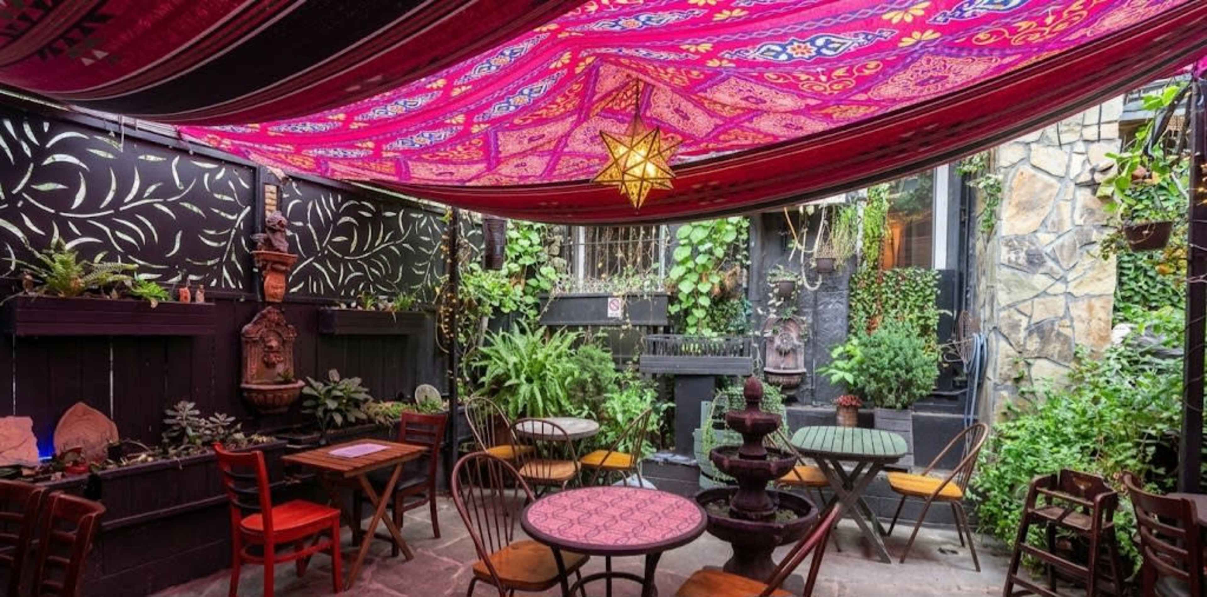A vibrant courtyard features colorful fabric draped overhead, surrounded by greenery and rustic furniture, with a decorative fountain in the center.