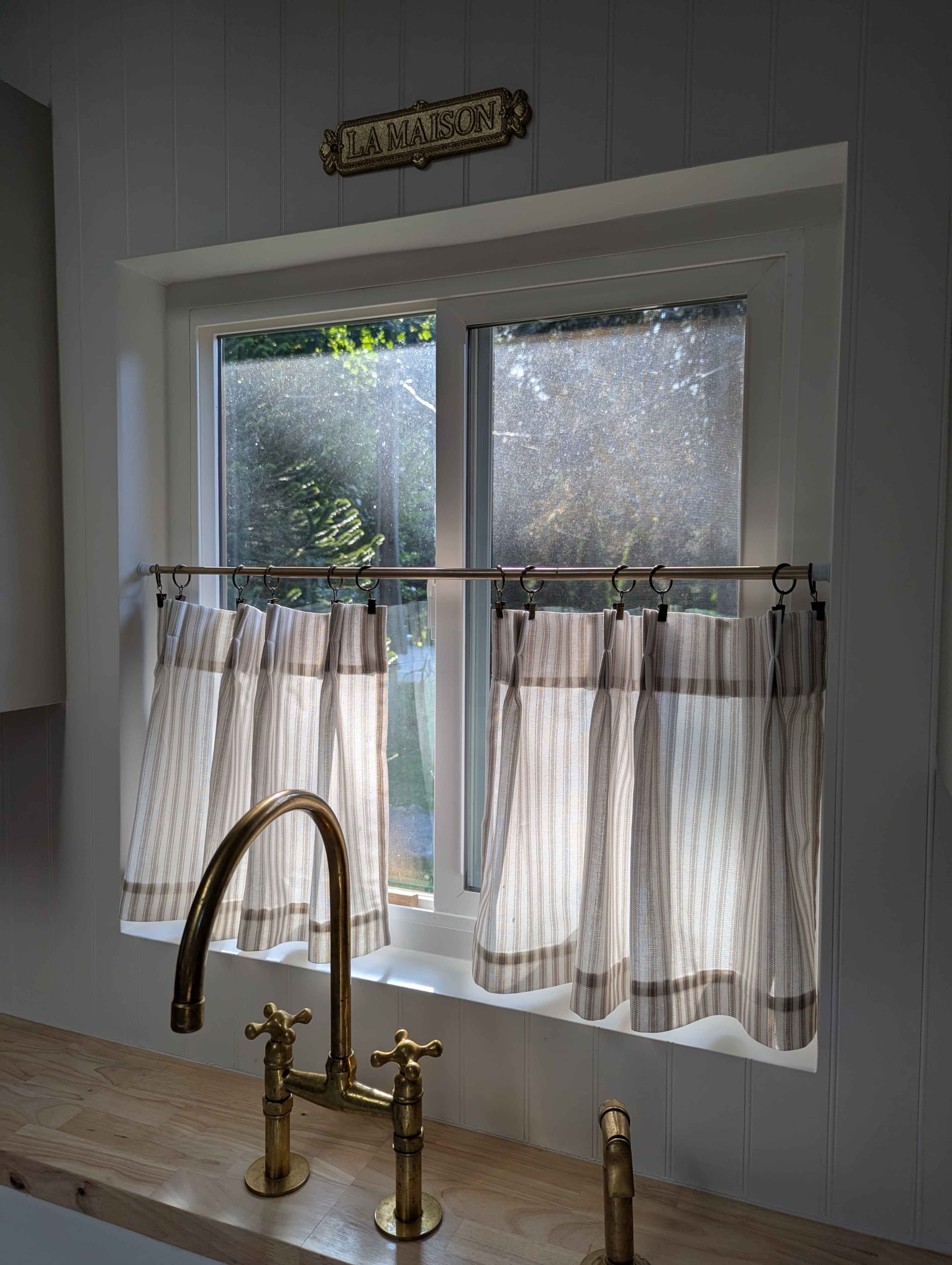 A kitchen window with striped sheer curtains and a wooden countertop beneath a brass faucet.