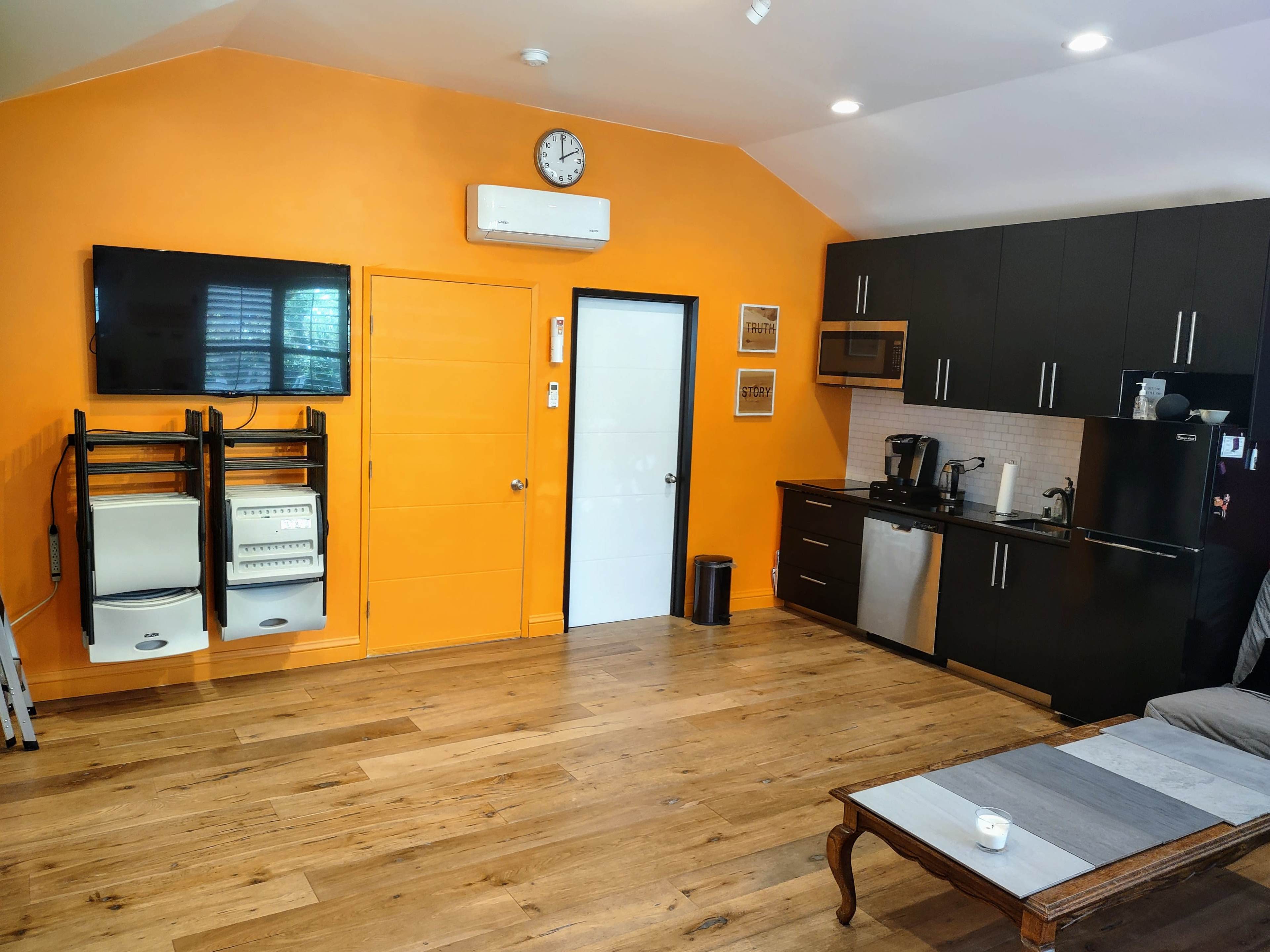 A modern kitchen and living area with an orange wall, black cabinets, a television mounted on the wall, and wooden flooring.