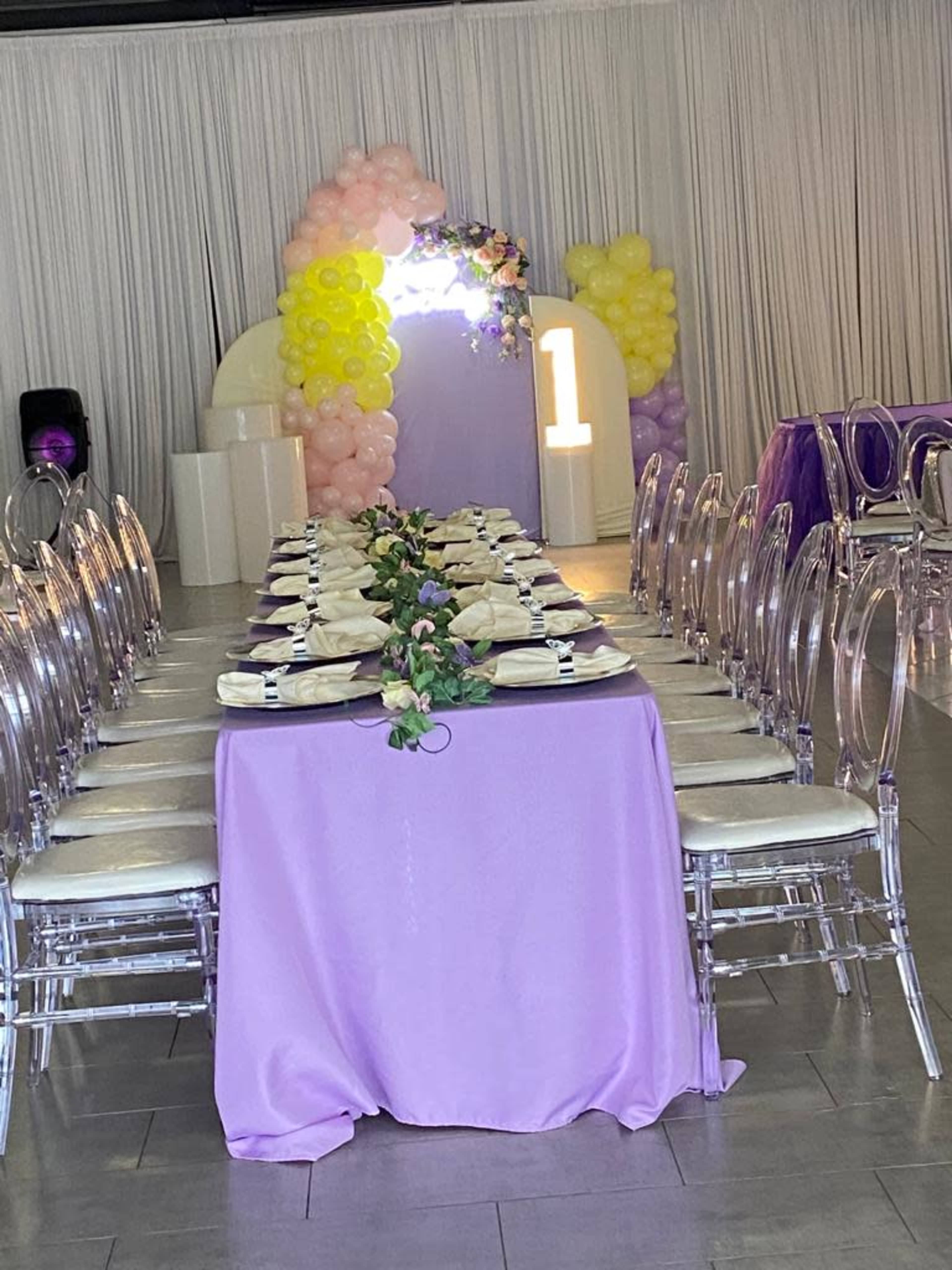 A long banquet table with purple tablecloths and white plates is set up in a decorated room featuring balloons and a large number one display at the back.