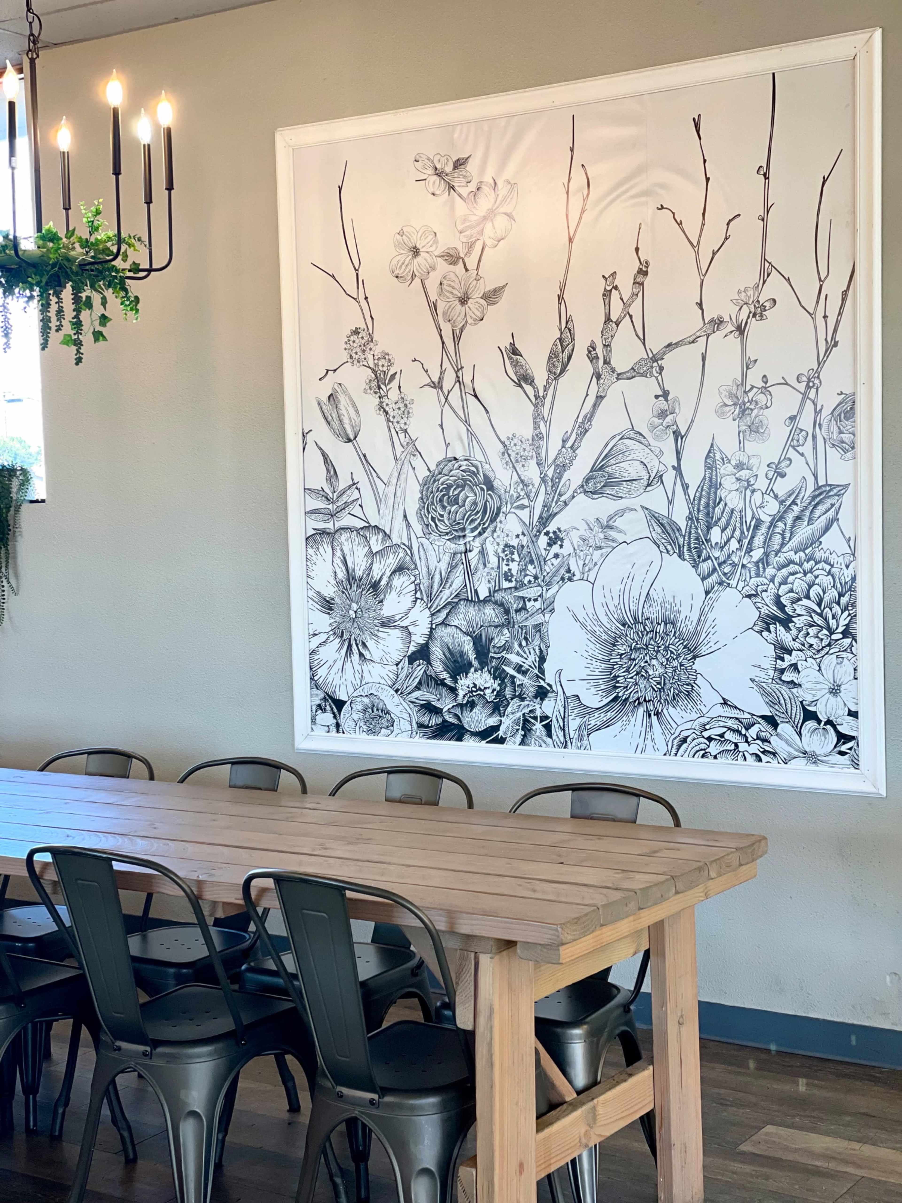The image shows a wooden dining table with metal chairs in a room featuring a large floral artwork on the wall.