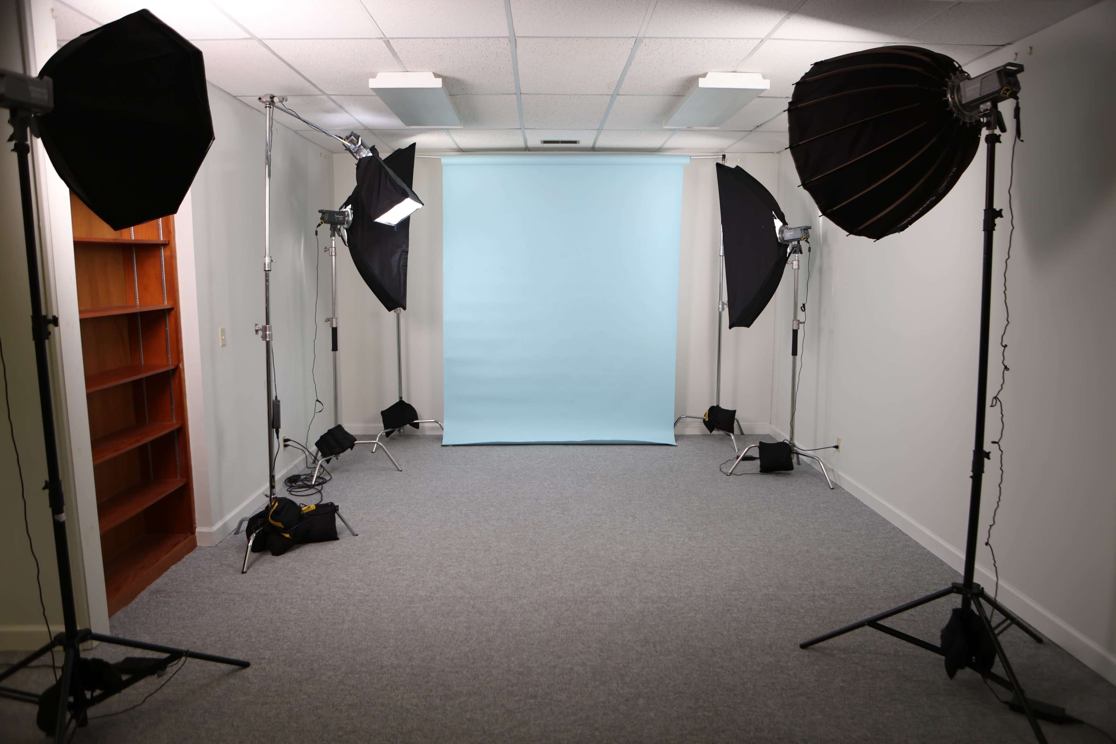 The image shows a photography studio with two softbox lighting setups, a light blue backdrop, and a carpeted floor.