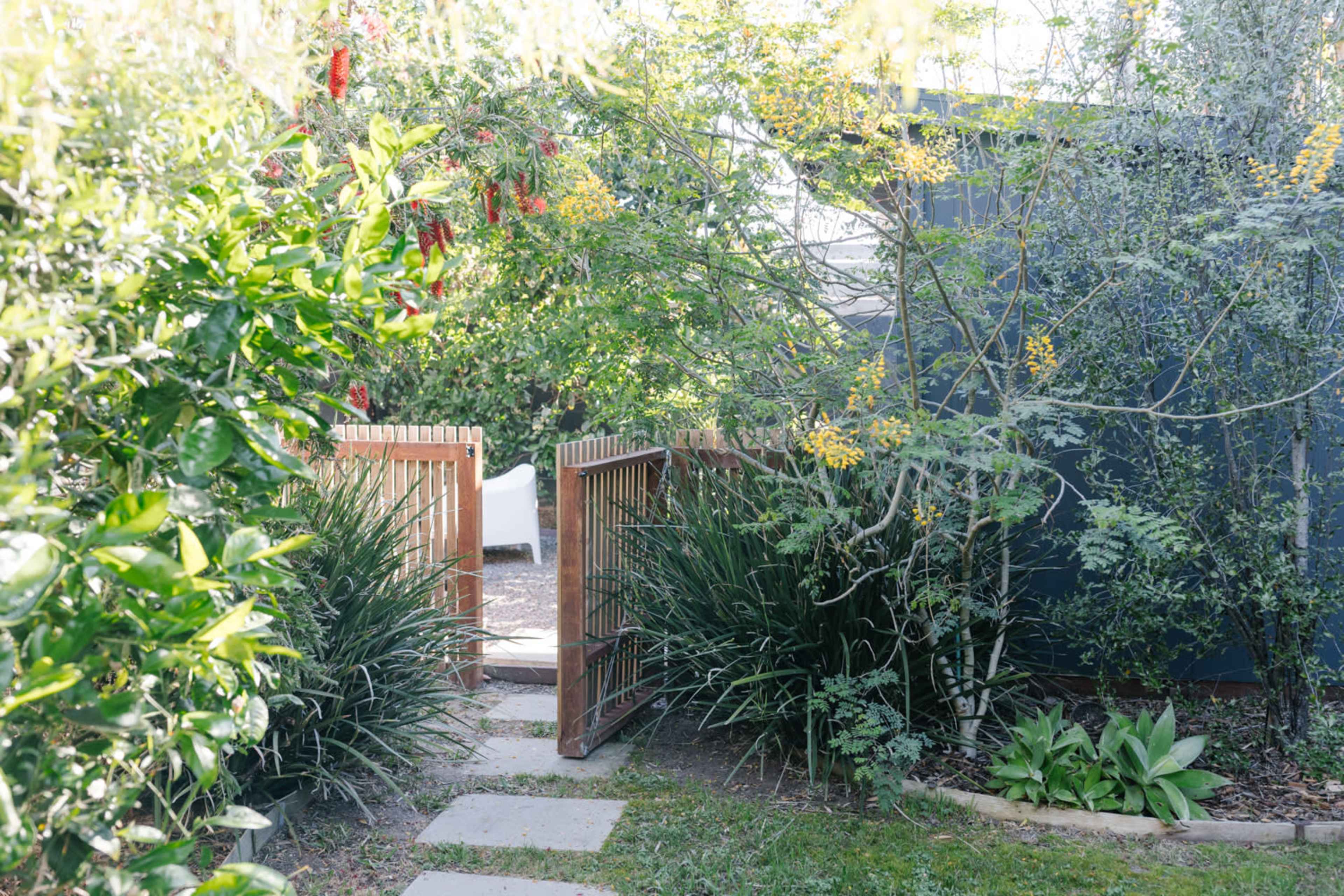 A wooden gate opens into a garden path surrounded by greenery and flowering plants.
