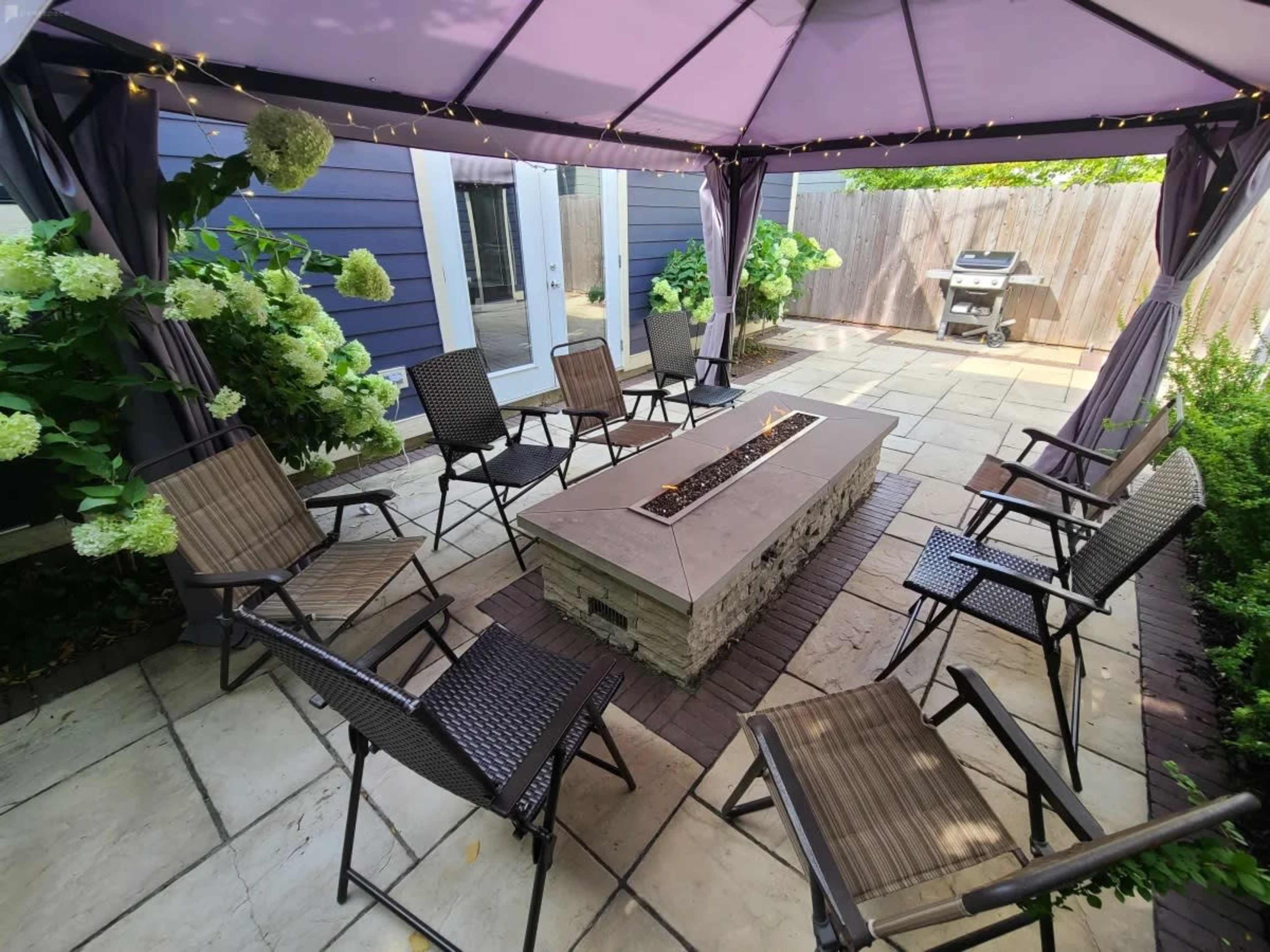 A patio area features a fire pit surrounded by several chairs under a canopy, with greenery and a grill in the background.
