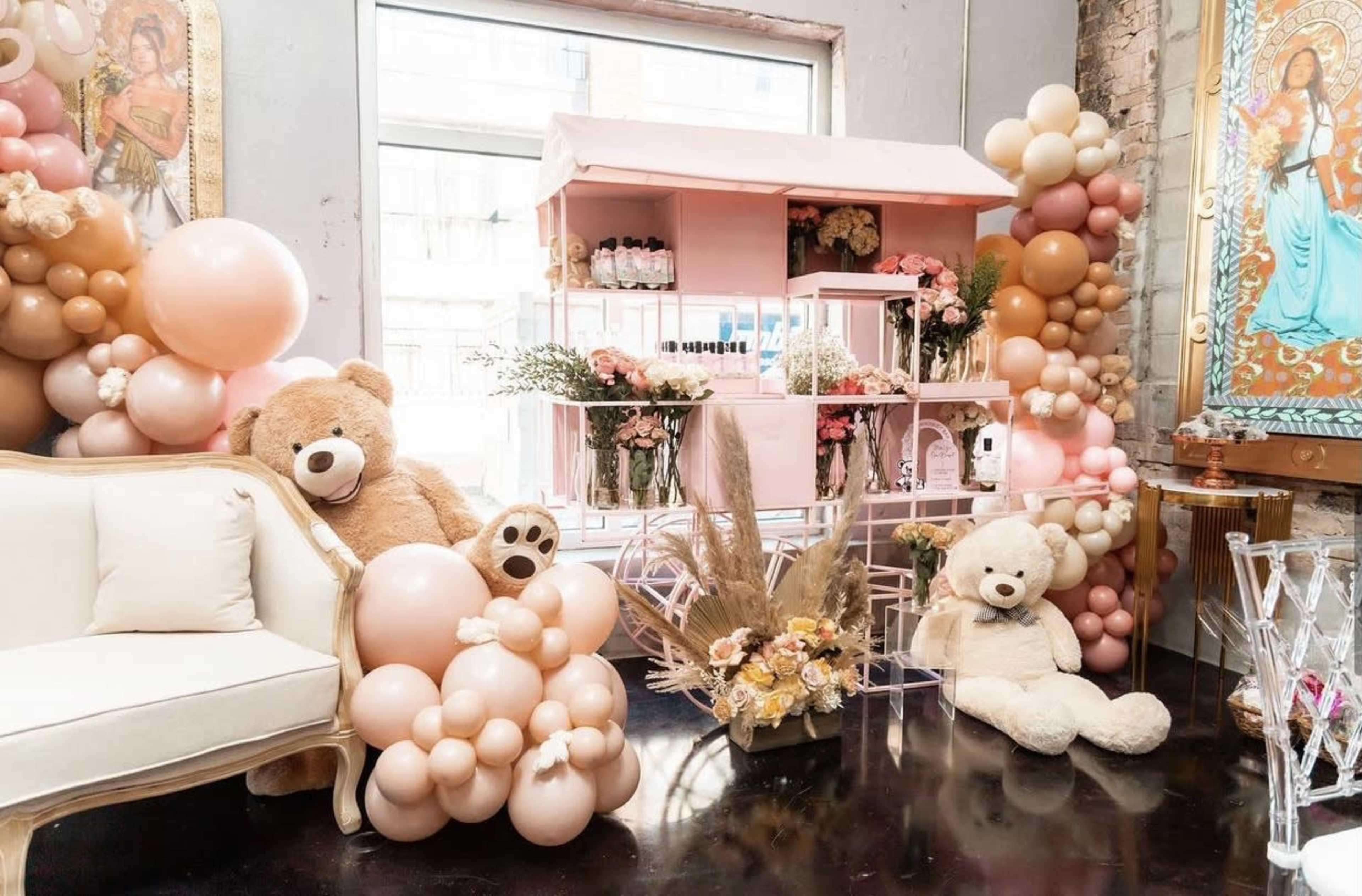 The scene features a cozy room adorned with a pink display stand filled with products, large teddy bears, and clusters of balloons in soft pastel colors.