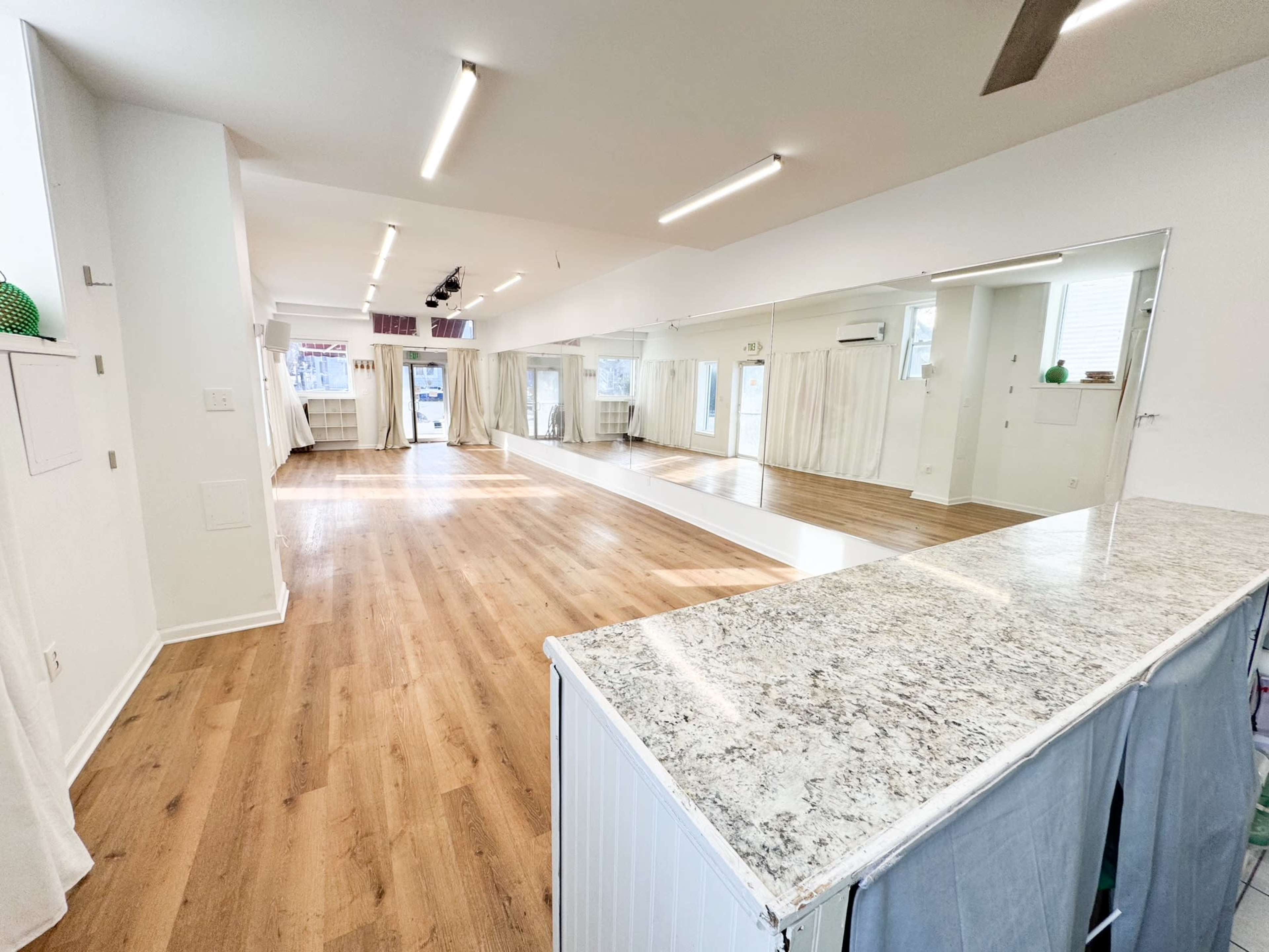 The image shows a spacious, brightly lit interior with wooden floors, a large mirror on one wall, and a countertop area at the forefront.