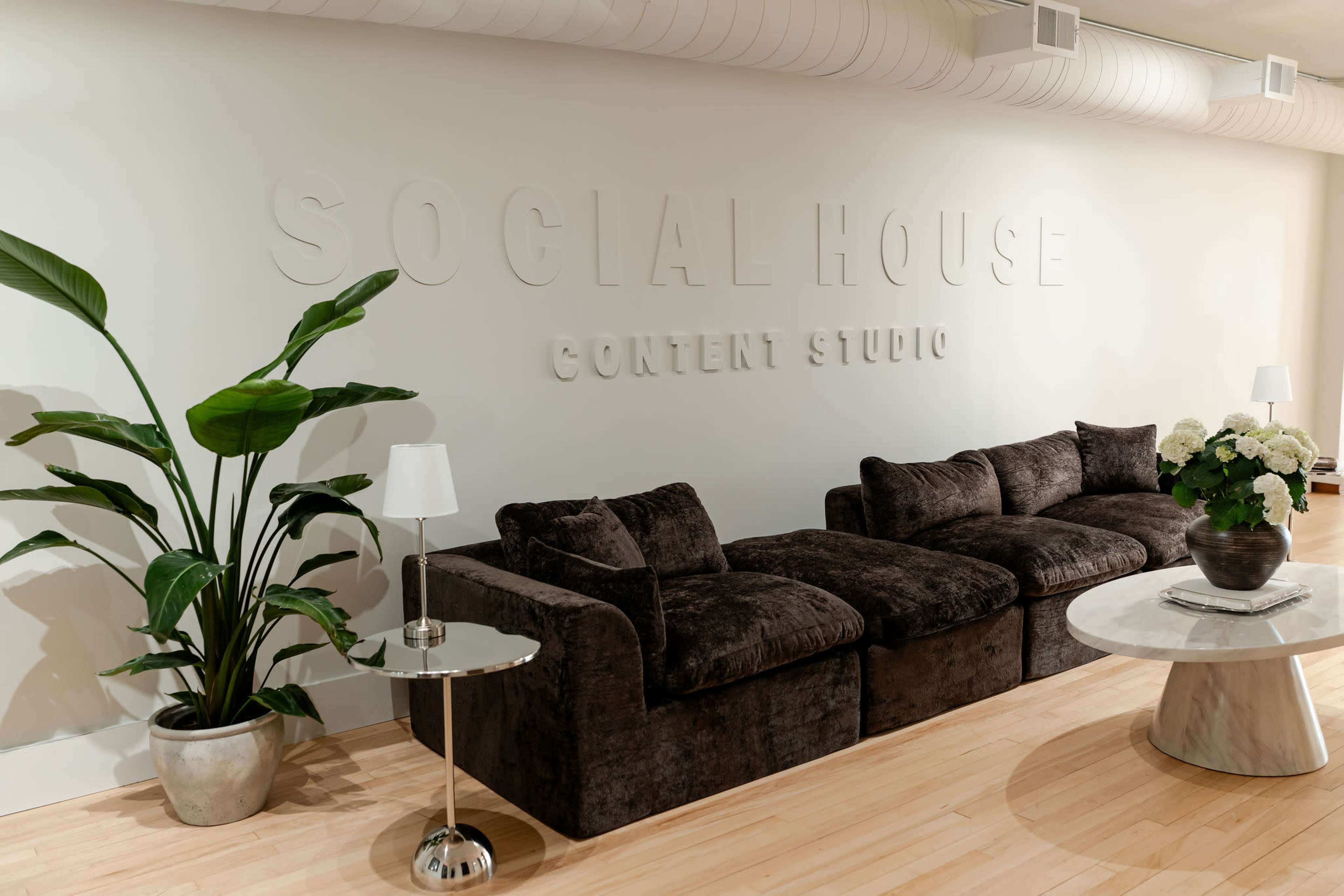 The image shows a modern interior with a dark, plush sofa facing a white wall that features the words "SOCIAL HOUSE CONTENT STUDIO," along with a small table, a plant, and a bouquet of flowers.