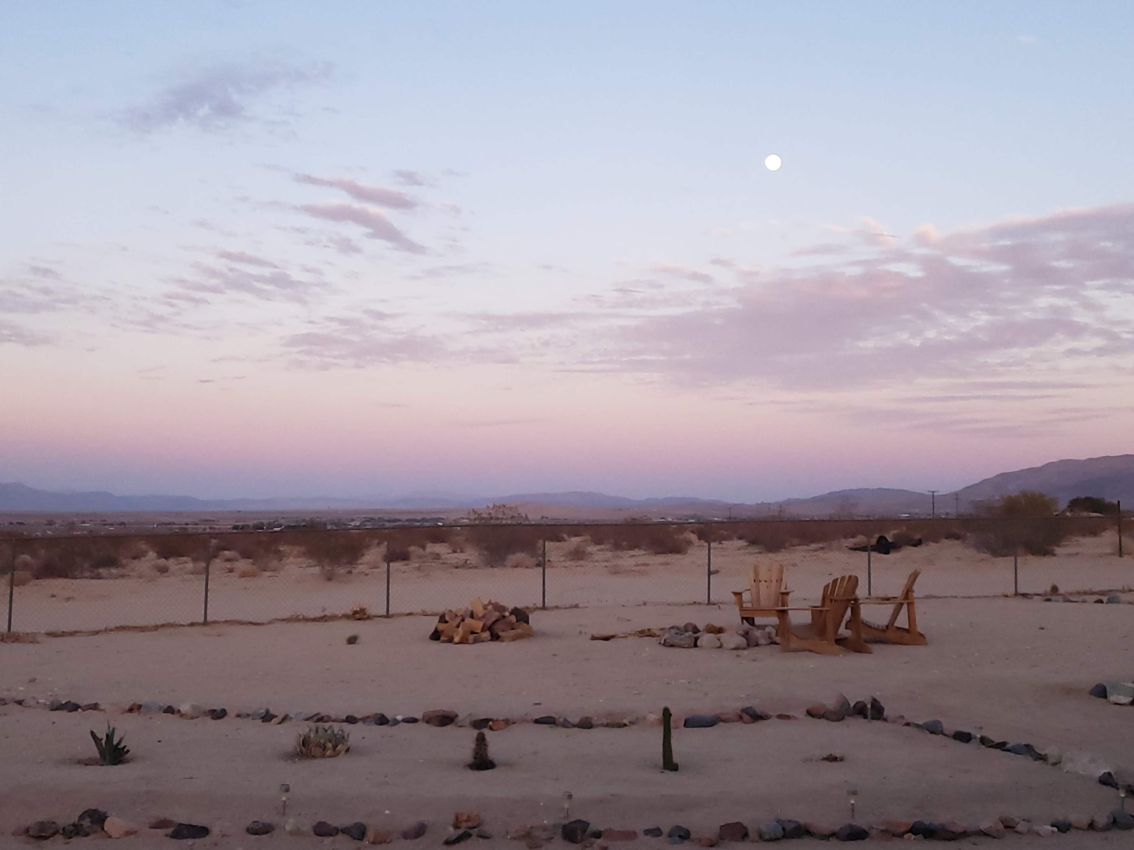 The scene shows a desert landscape at dusk with a fire pit surrounded by stones and wooden chairs facing an expansive view of mountains under a pale moon.