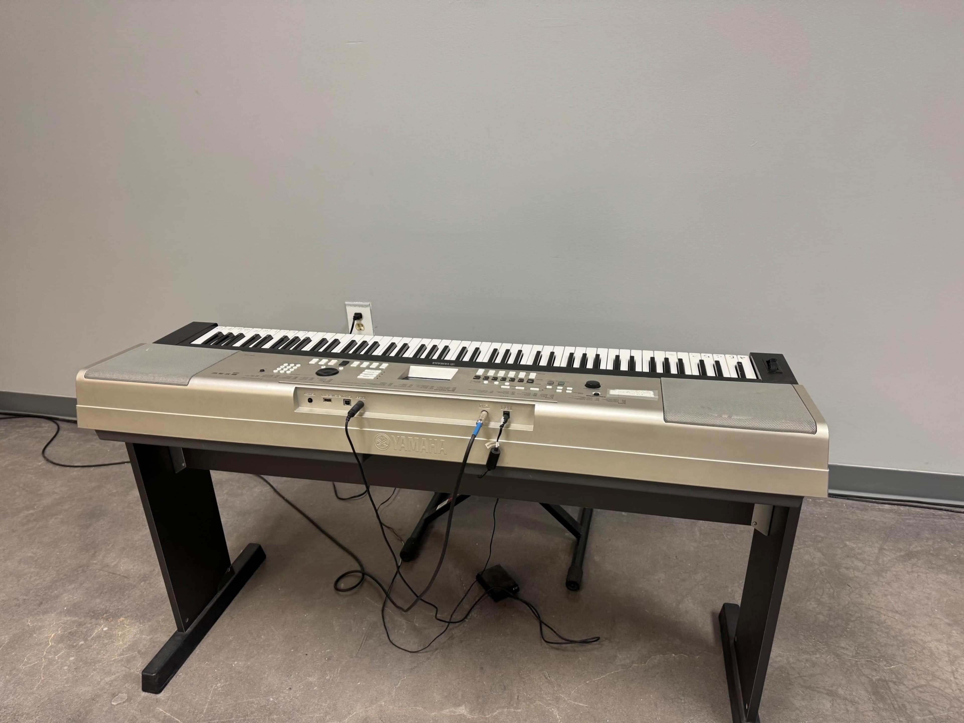The image shows a silver electronic keyboard placed on a black stand against a plain gray wall.