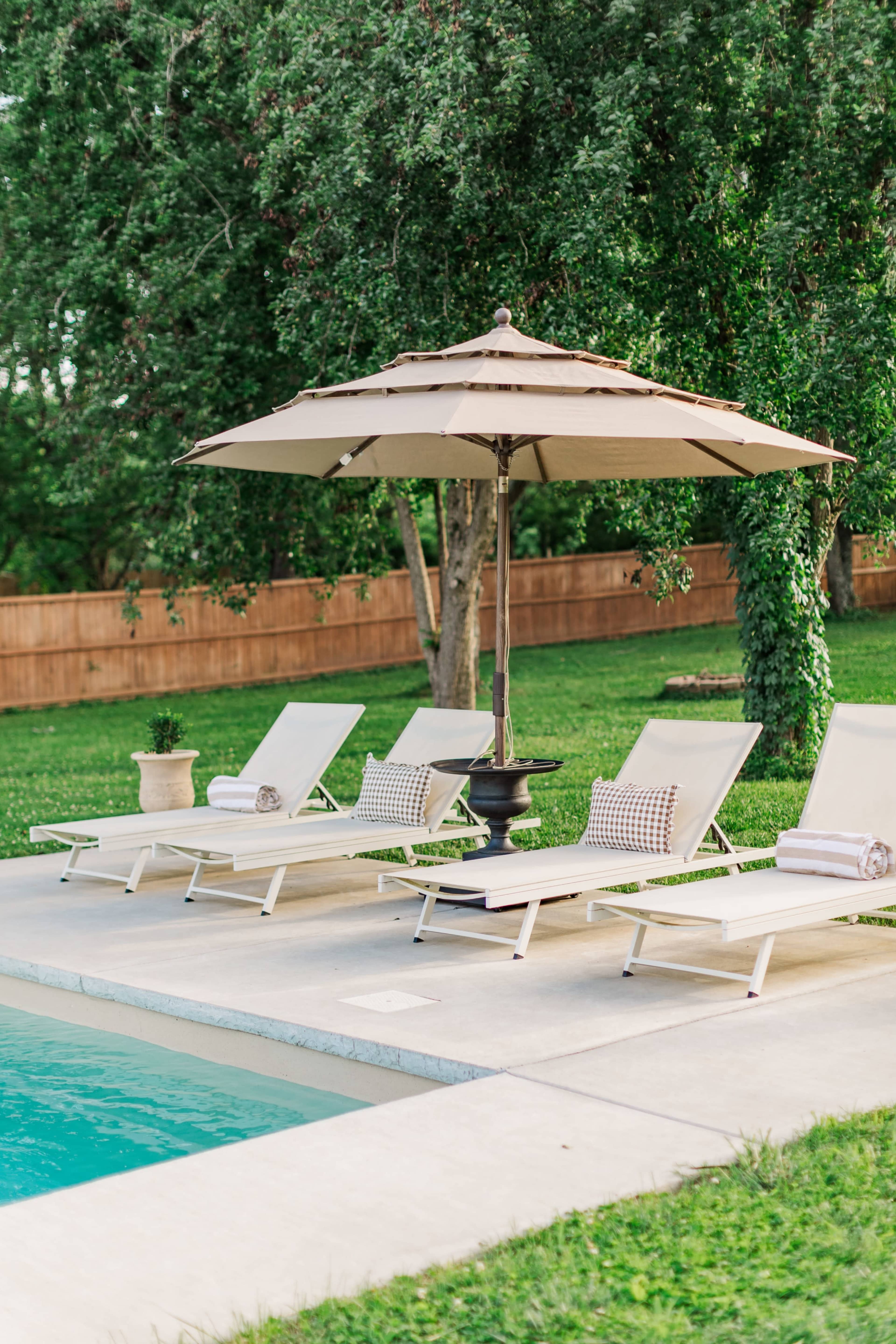 The image depicts a serene outdoor pool area with four lounge chairs under a large umbrella, surrounded by green grass and trees.
