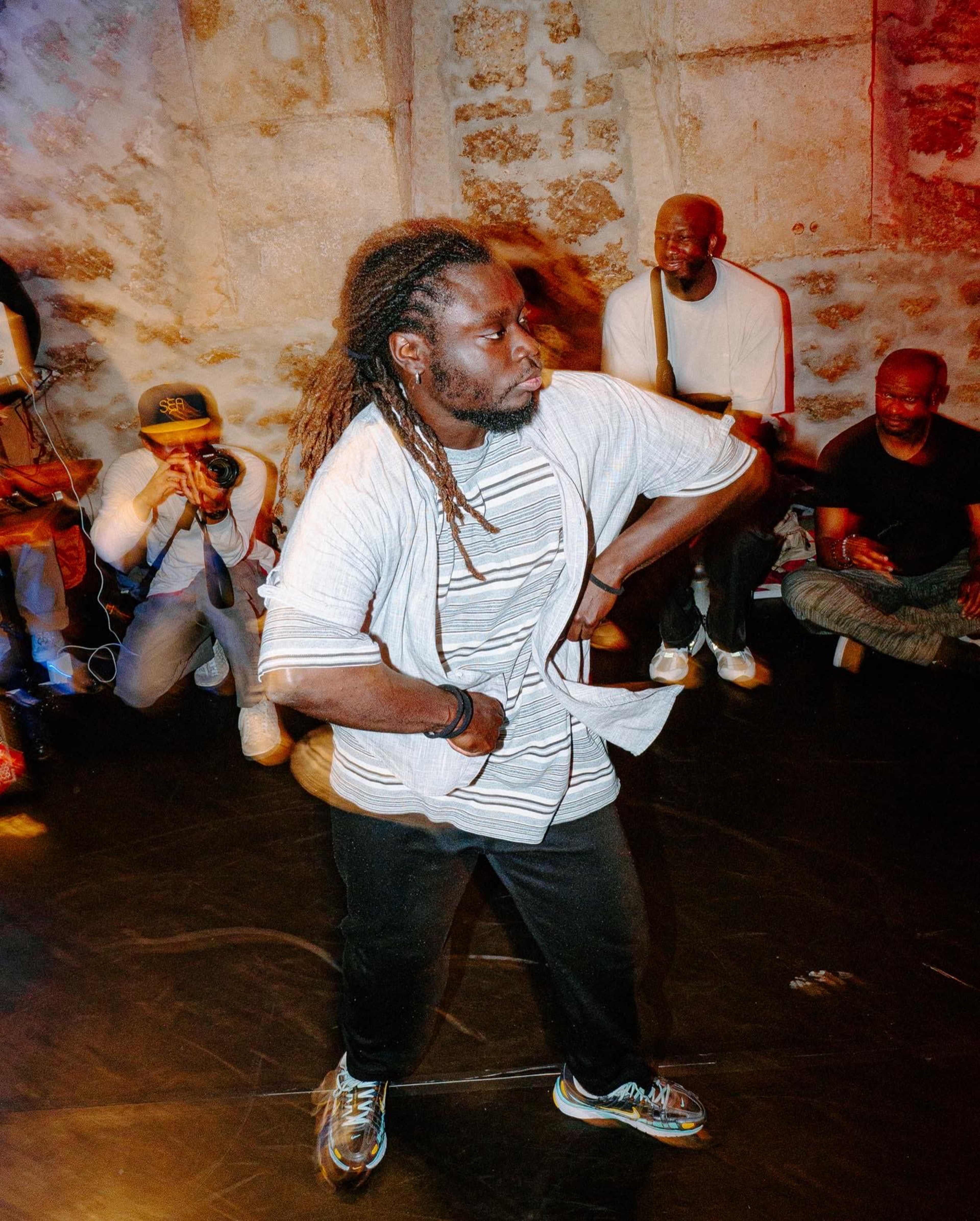 A dancer performs confidently on a dimly lit stage while photographers capture the moment and an audience watches nearby.