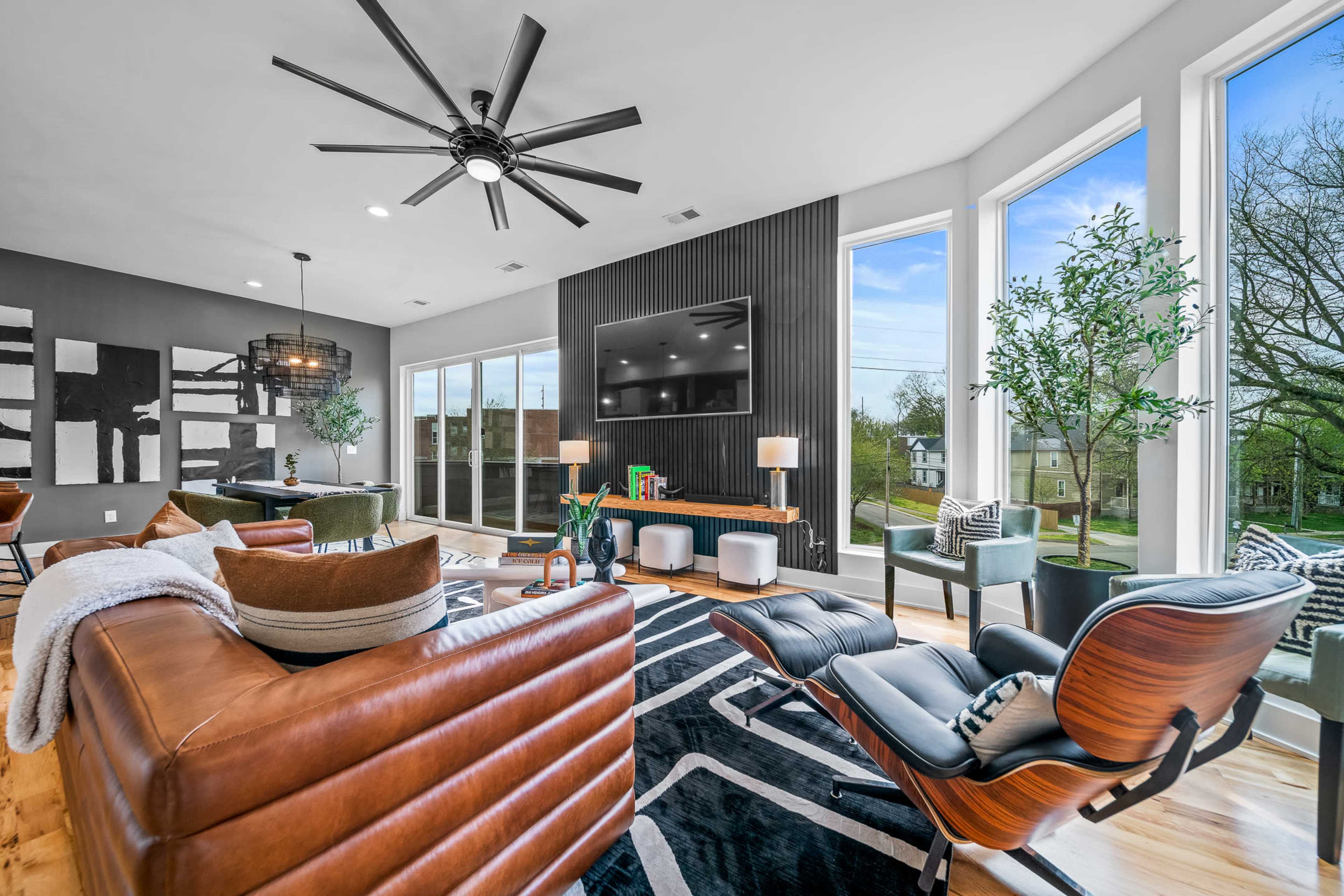 A modern living room features large windows, a flat-screen TV mounted on a black accent wall, and a mix of leather and wood furniture.