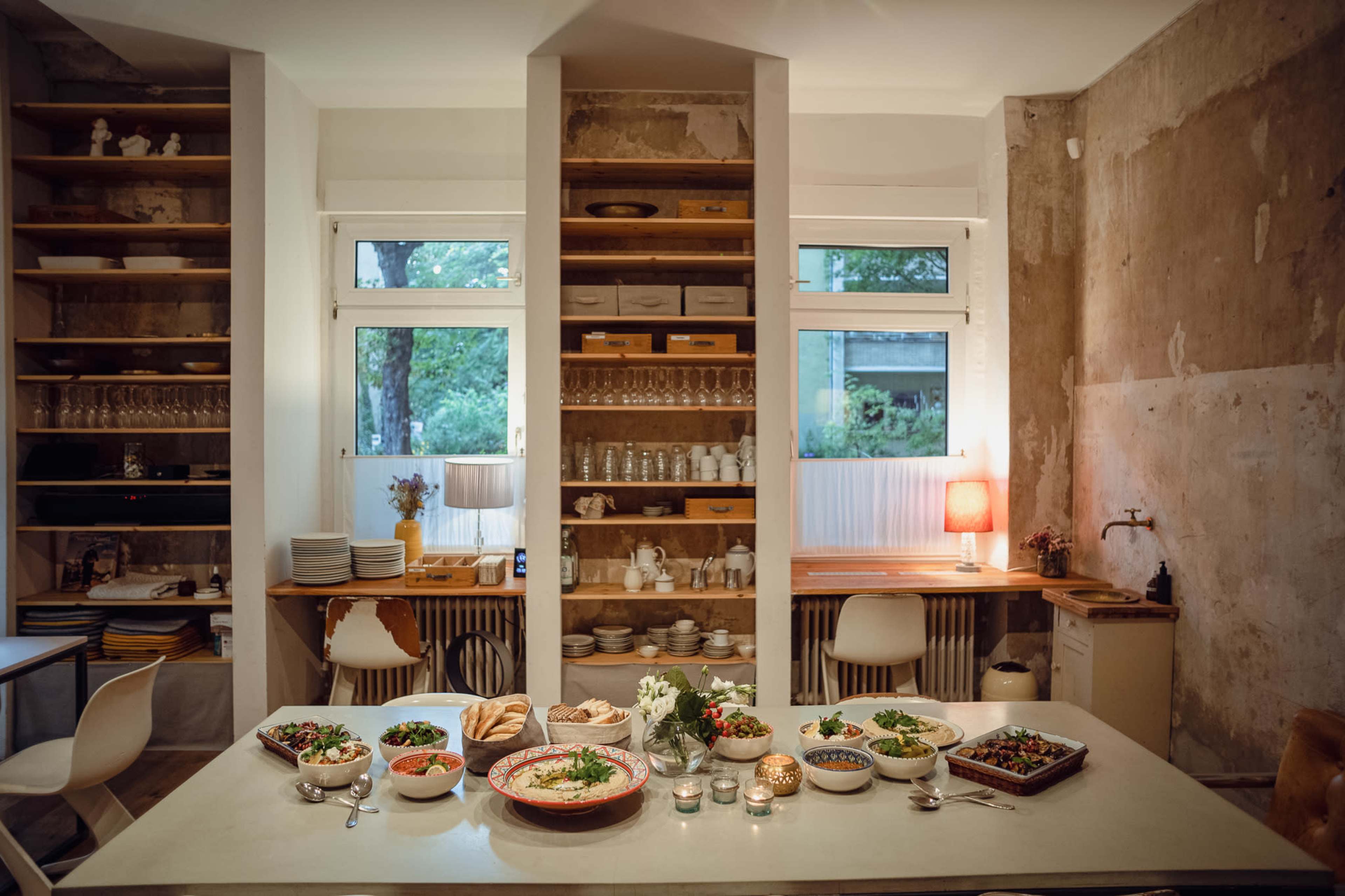 A dining table is set with various dishes and garnishes in a partially renovated kitchen featuring shelves filled with tableware and a view of greenery outside.