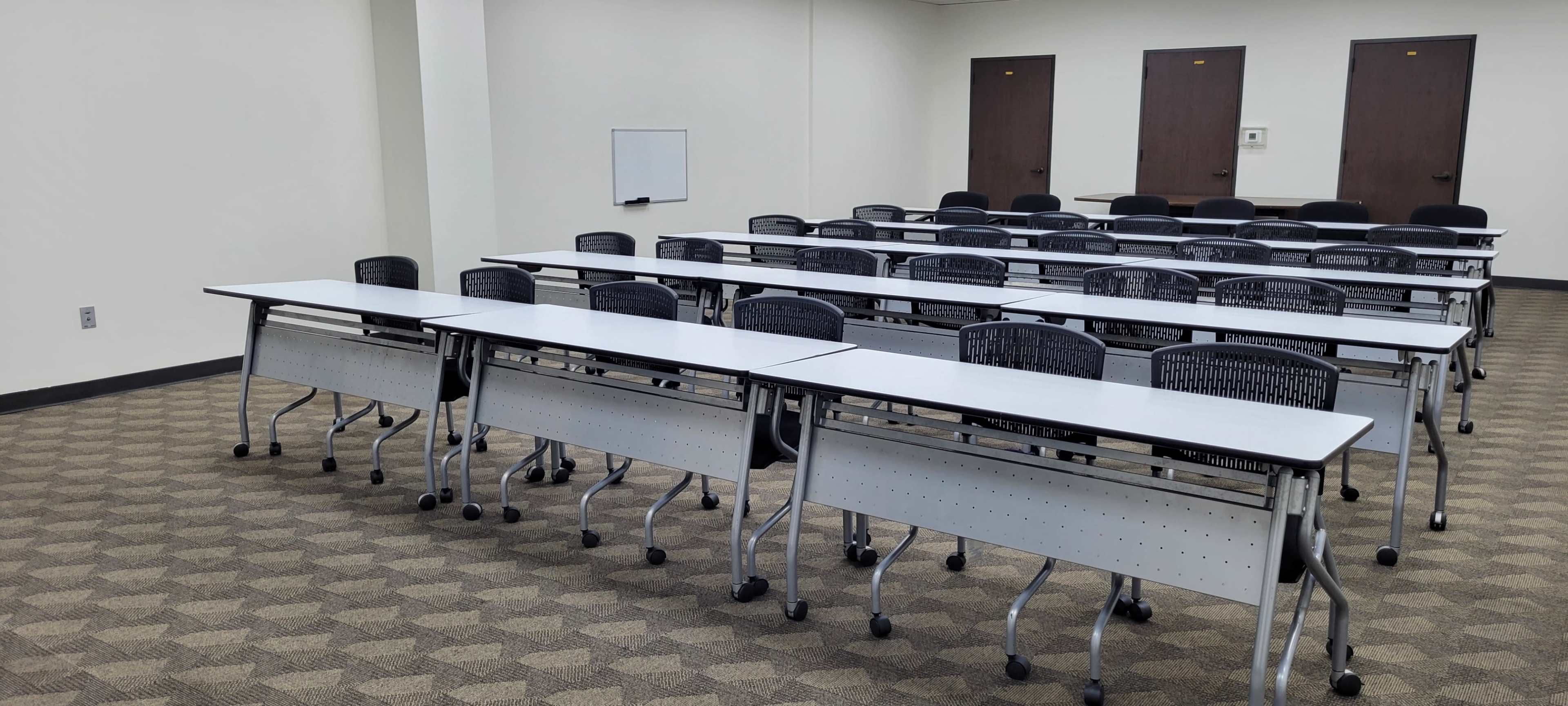 The image shows a classroom with multiple rows of tables and chairs arranged in a neat layout, featuring a whiteboard on one wall and closed doors in the background.