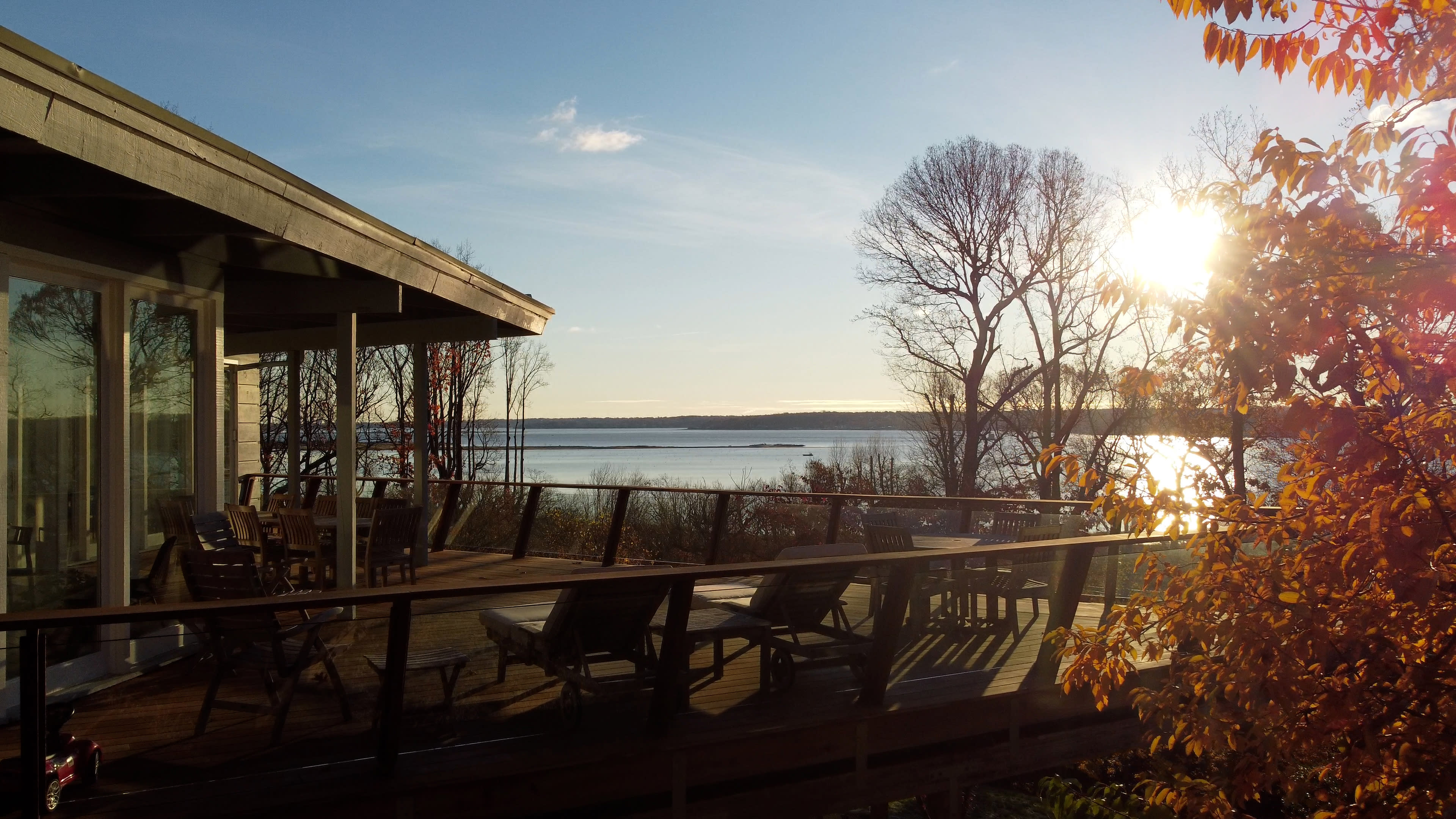 A wooden deck with chairs overlooks a calm body of water under a clear sky, with sunlight streaming through trees.
