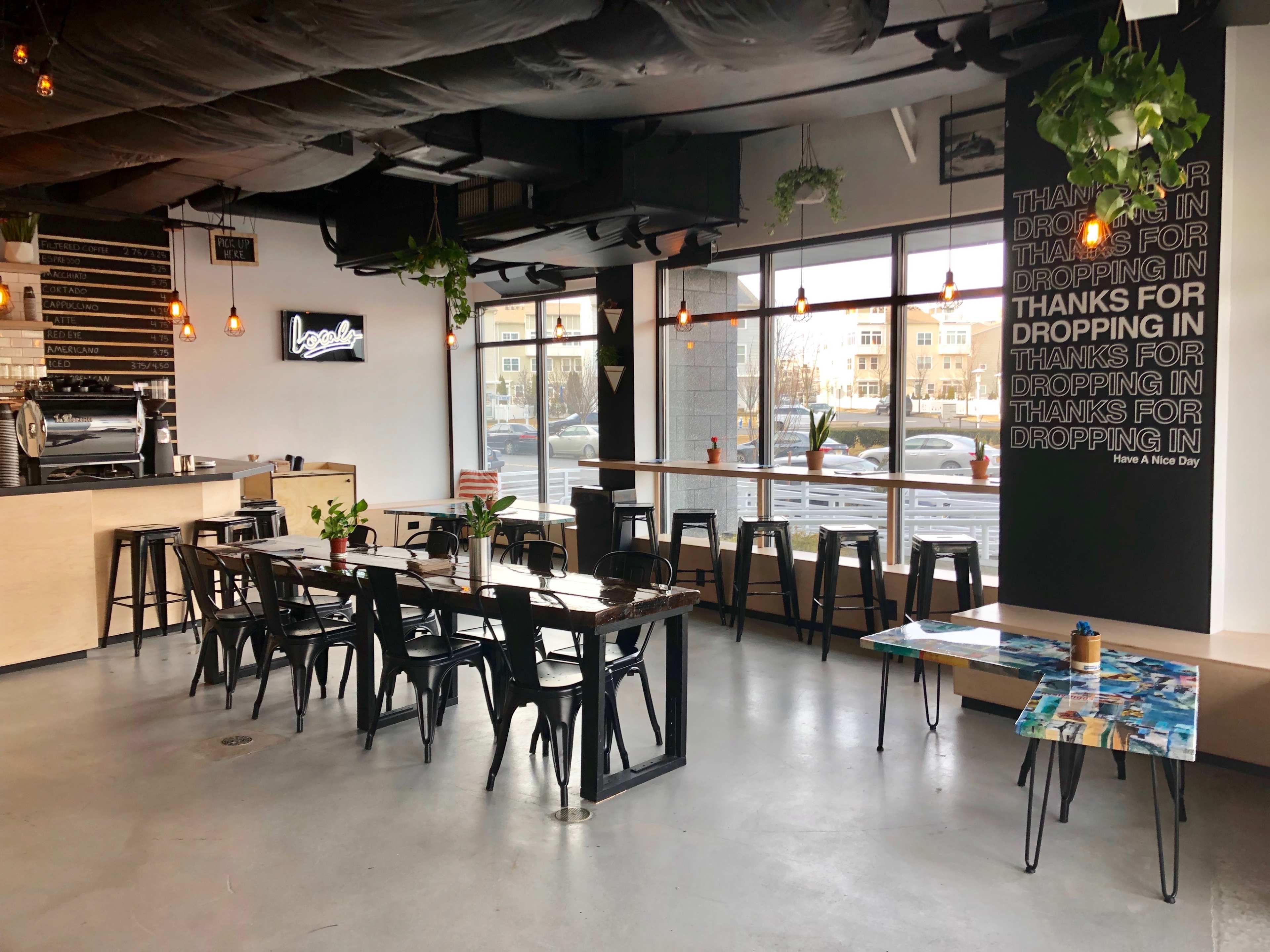 The image shows a modern café interior with wooden tables, black metal chairs, and large windows letting in natural light, featuring a coffee bar and decorative plants.