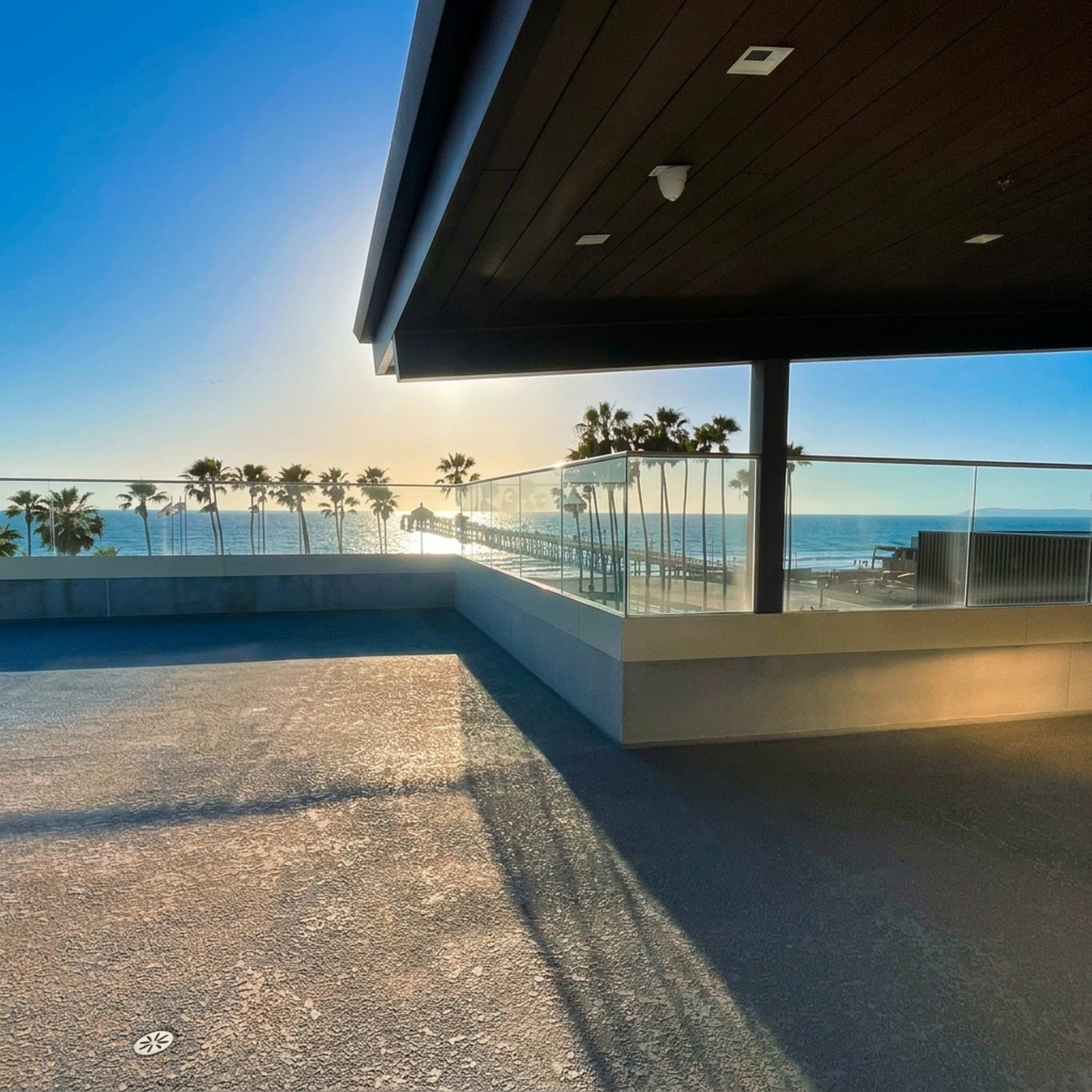 A modern outdoor terrace overlooks a serene beach, framed by palm trees and sunlight reflecting off the ocean.
