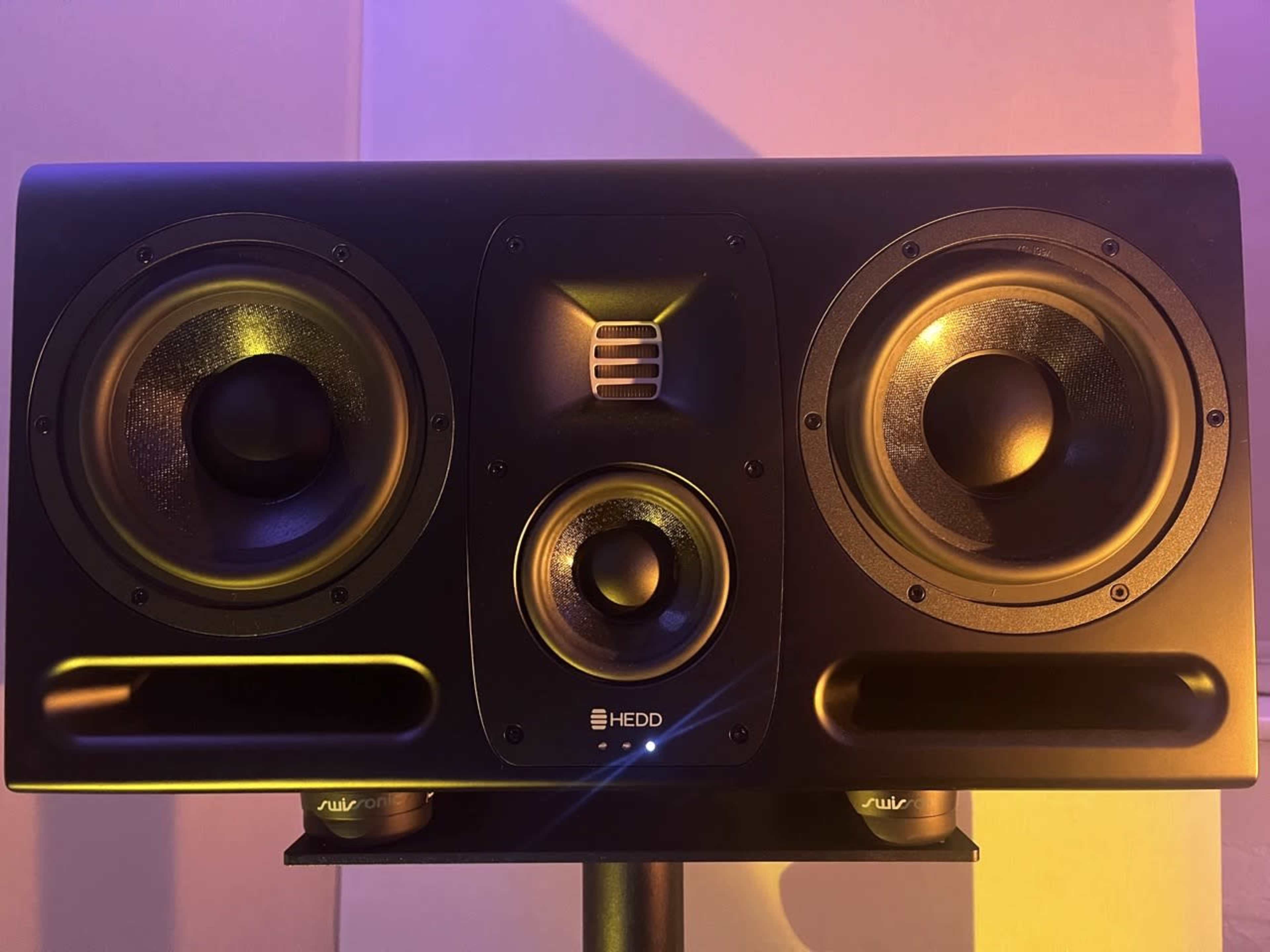 The image shows a large studio monitor speaker with three visible drivers and a central logo on a black casing.