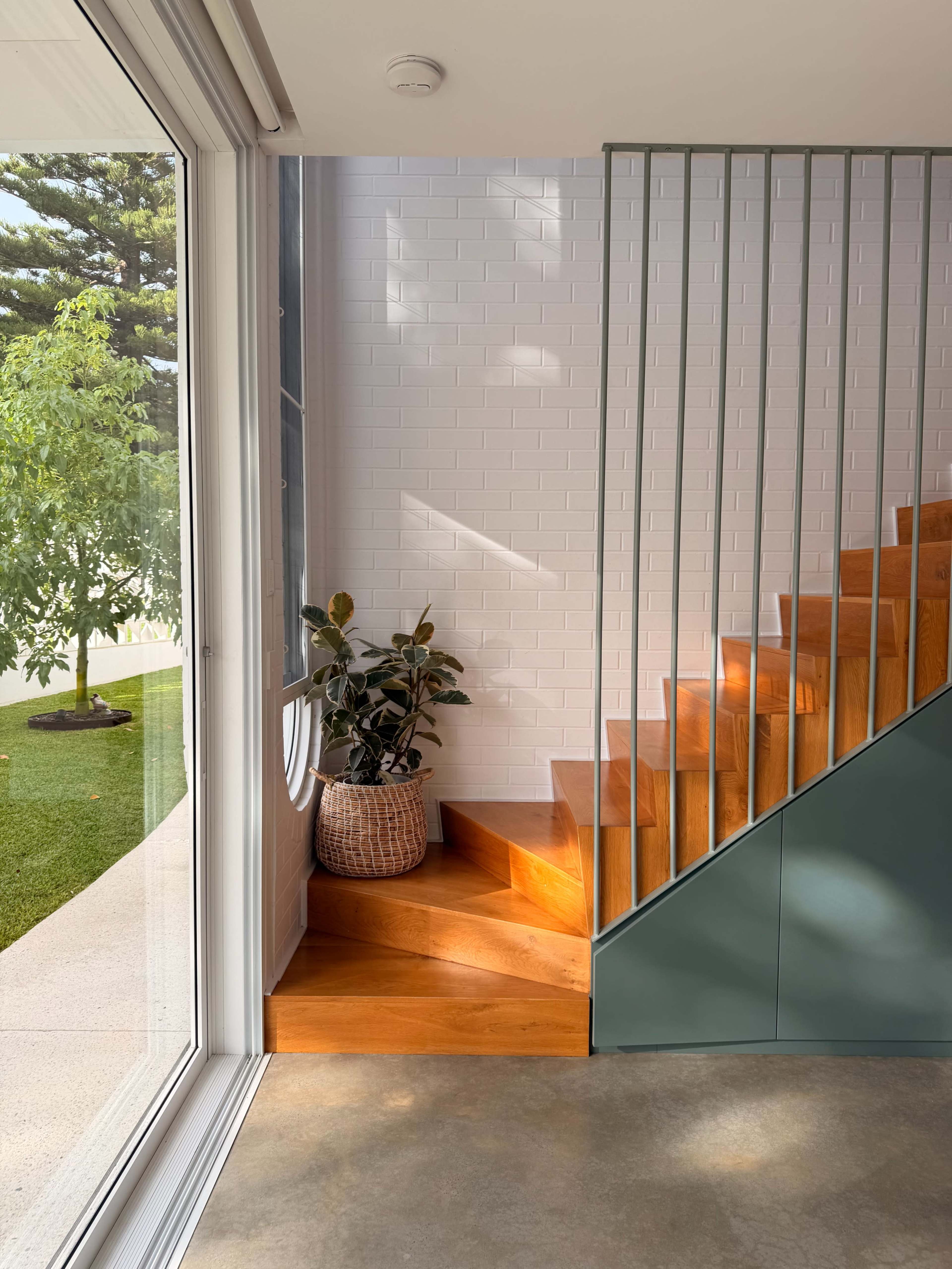 A wooden staircase with green railings ascends beside a potted plant in a well-lit entryway featuring large windows.