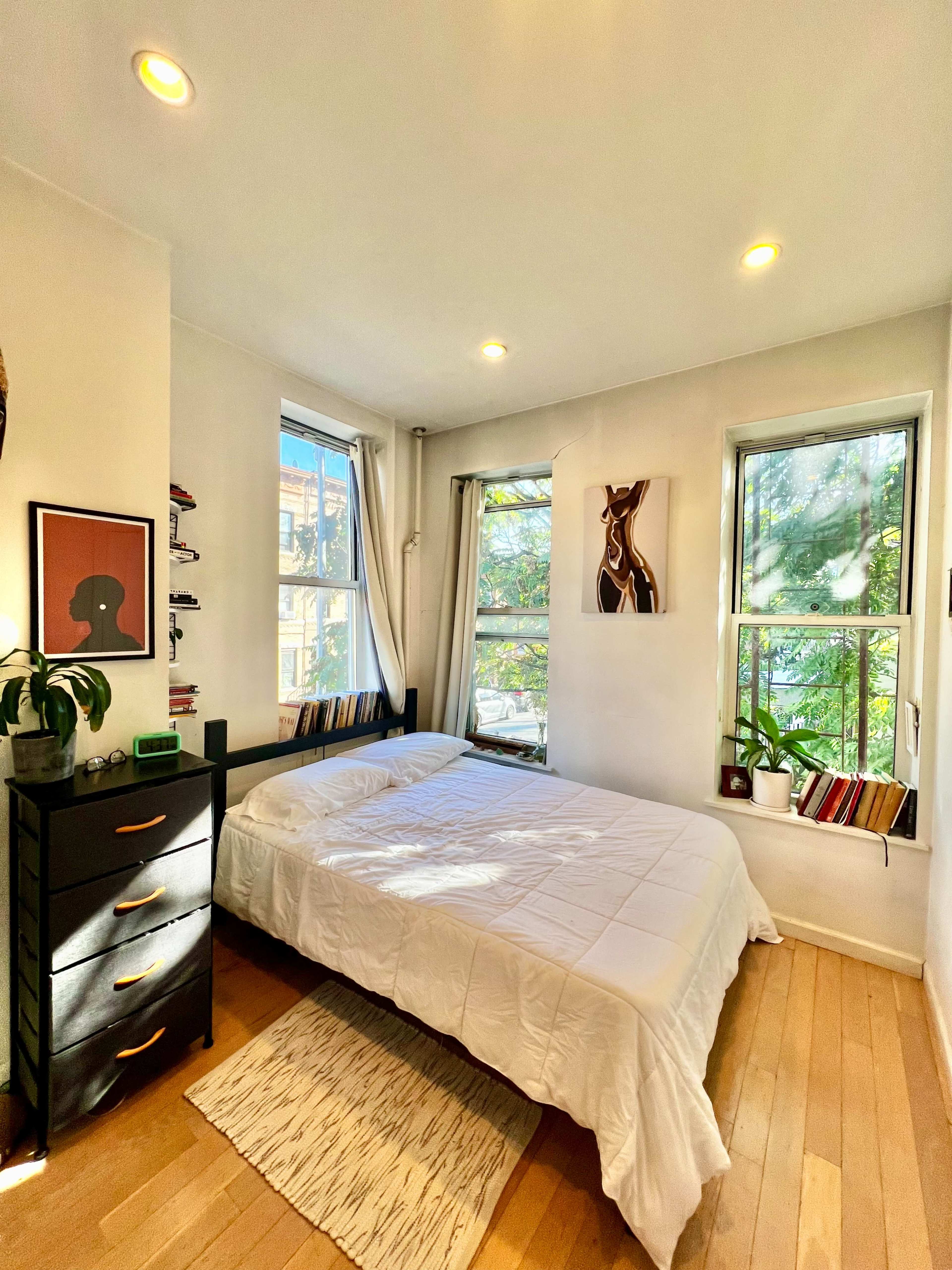 The image shows a small, well-lit bedroom featuring a bed, a black dresser, and two windows with greenery visible outside.