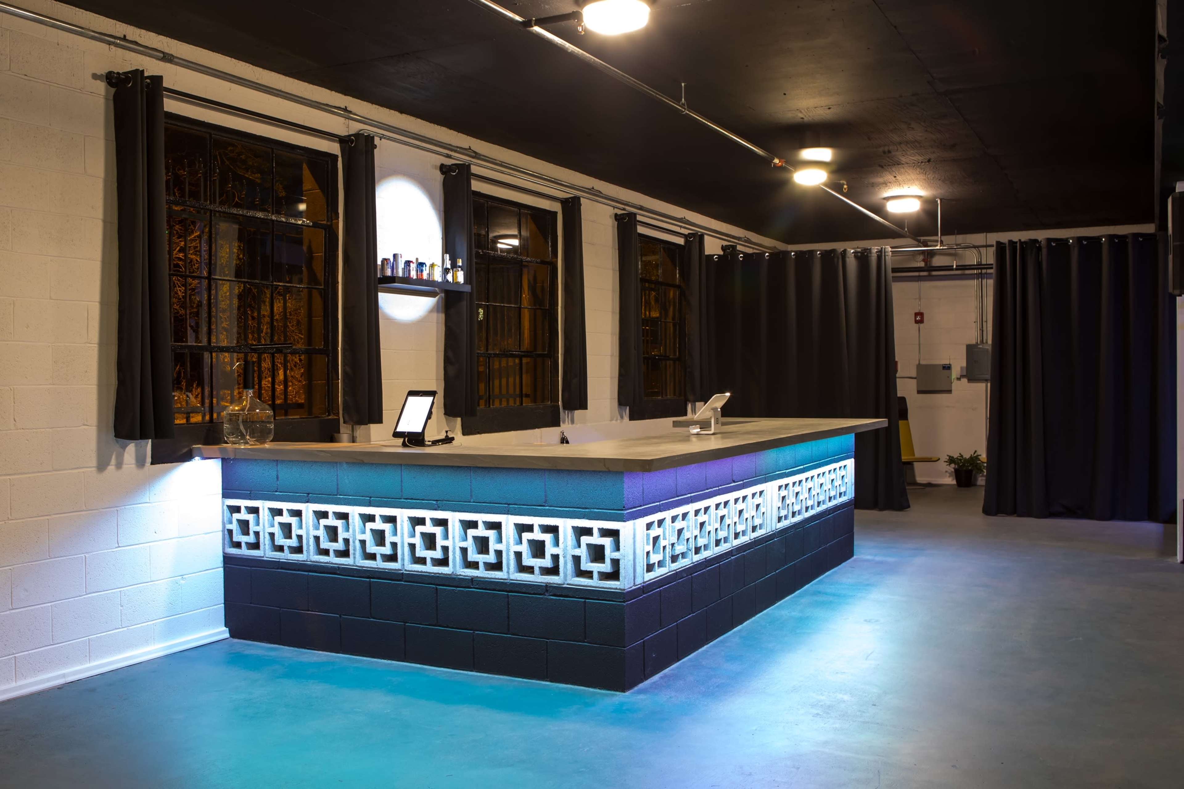 A modern bar area with a sleek counter illuminated by blue lighting, surrounded by dark curtains and large windows.
