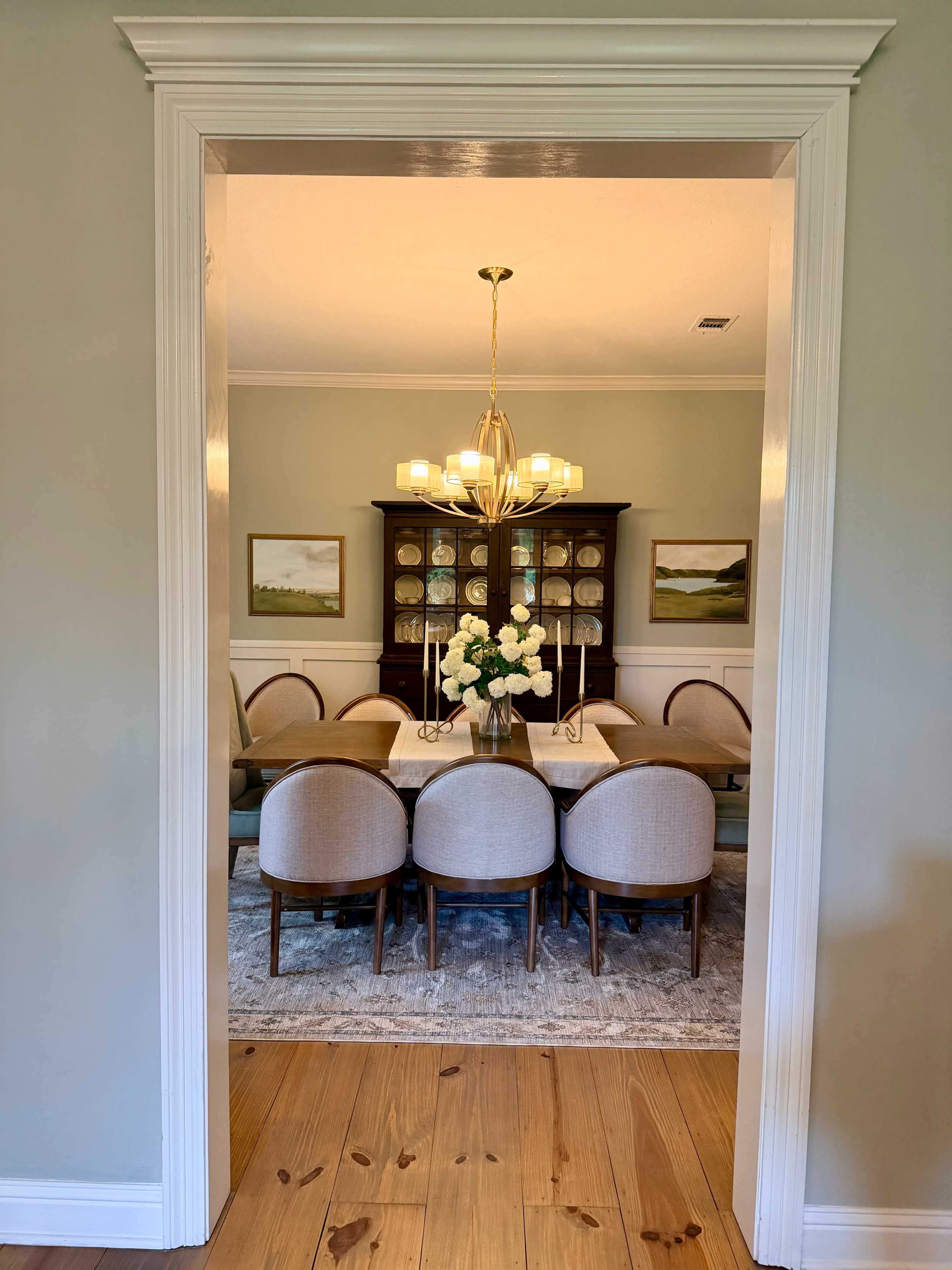 A well-lit dining room features a long table surrounded by upholstered chairs, with a chandelier above and a floral centerpiece in the middle.