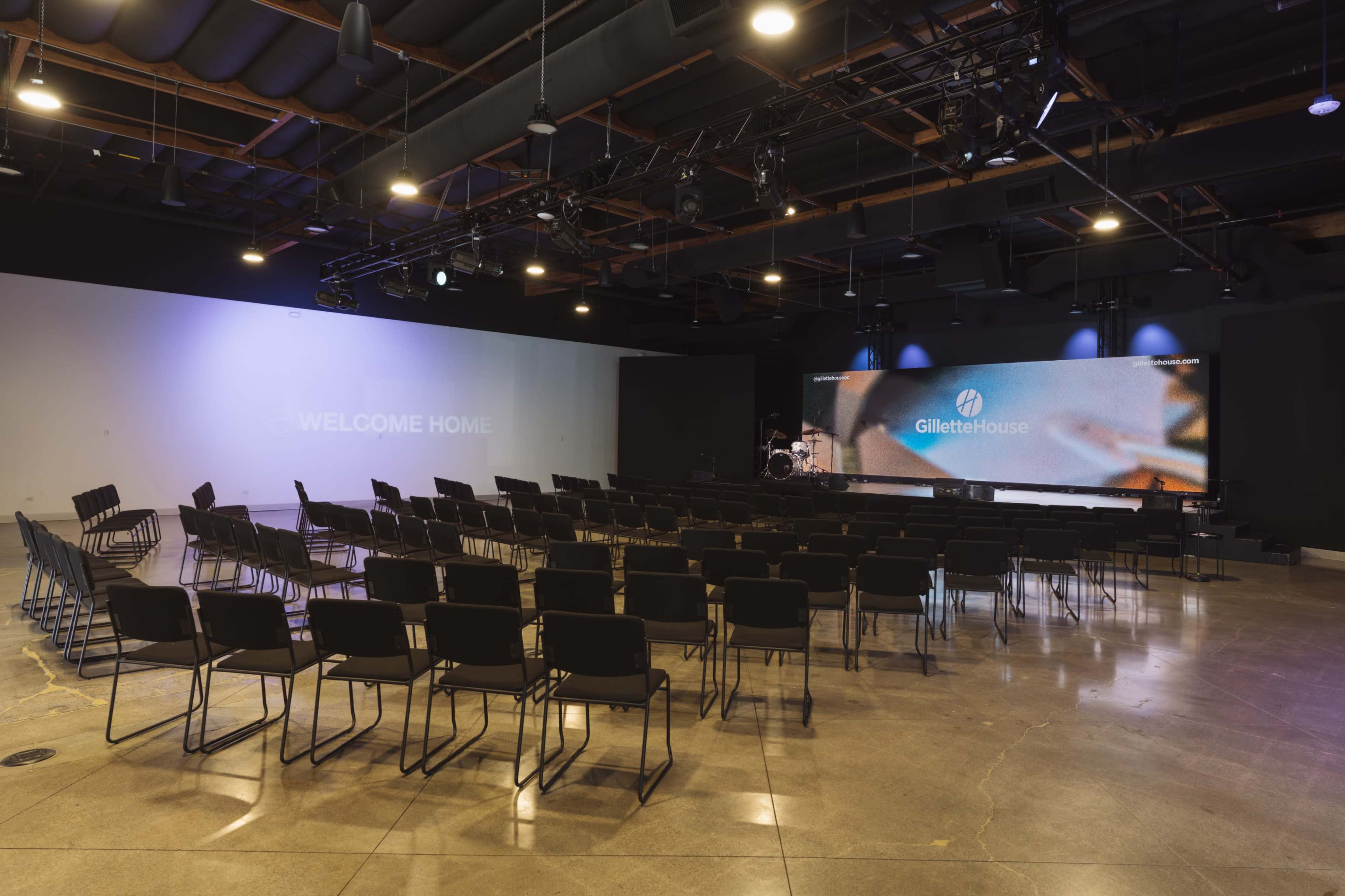The image shows a spacious indoor venue set up for an event, featuring rows of black chairs facing a large screen with "GilletteHouse" displayed.