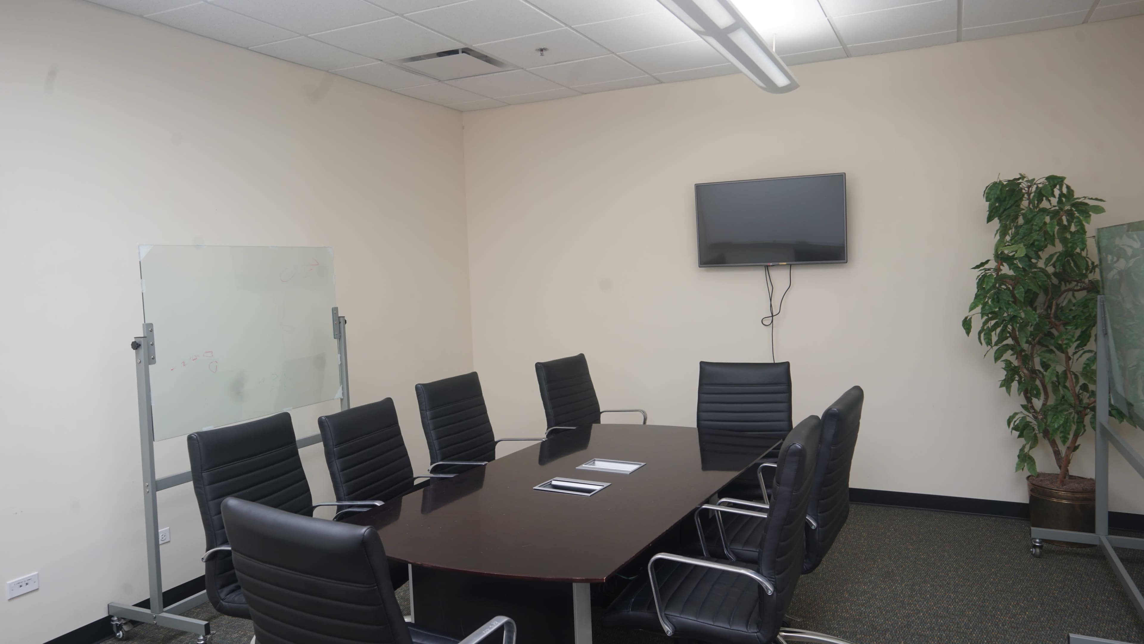 A conference room features a large rectangular table surrounded by black leather chairs, with a television mounted on the wall and a glass whiteboard.