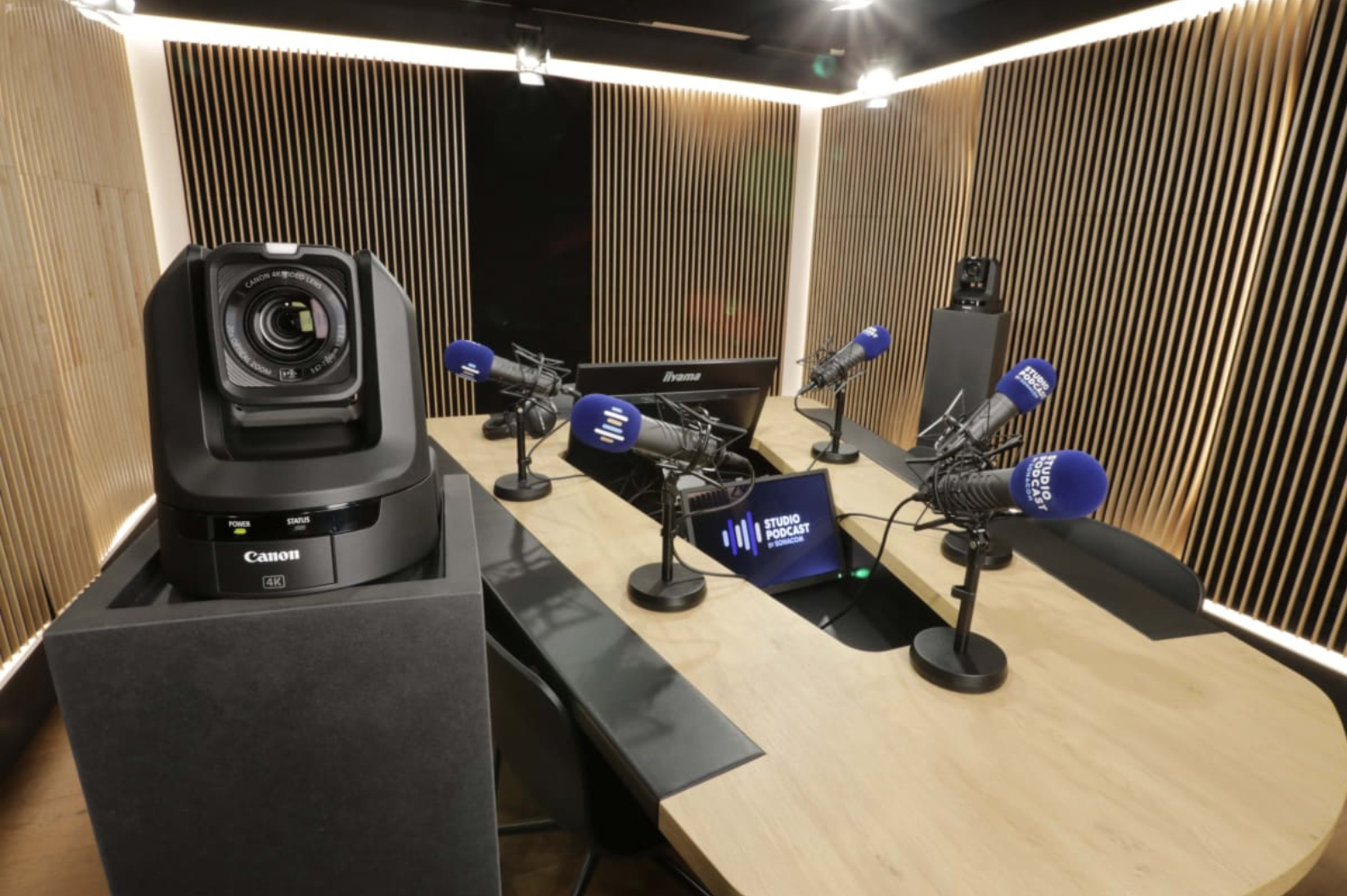 A modern recording studio with multiple microphones positioned at a central table, a camera on the left, and wooden slatted walls.