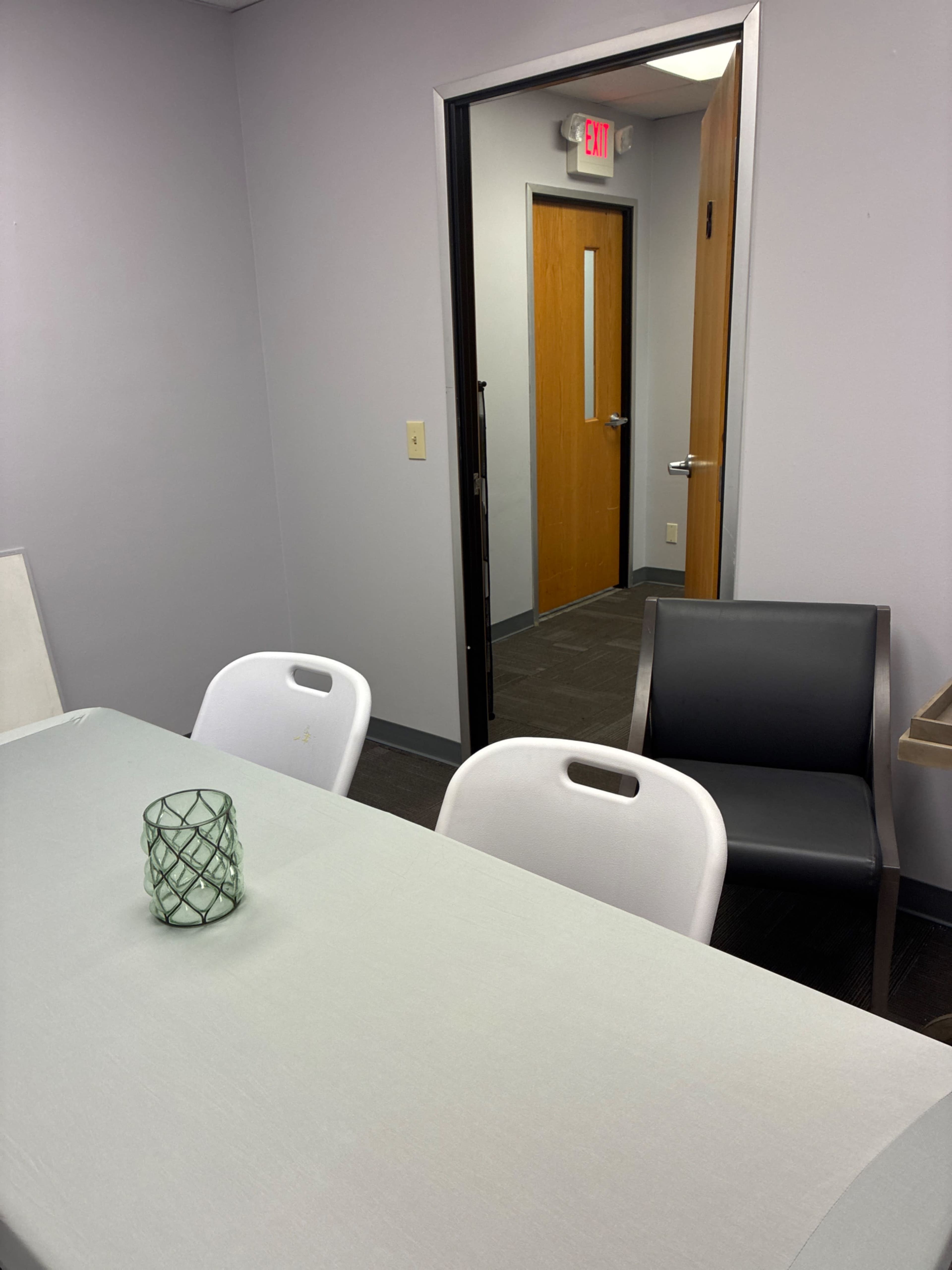 A gray meeting room features a table with a green decorative candle holder, a white chair, and a gray chair, with an open doorway leading to a corridor.