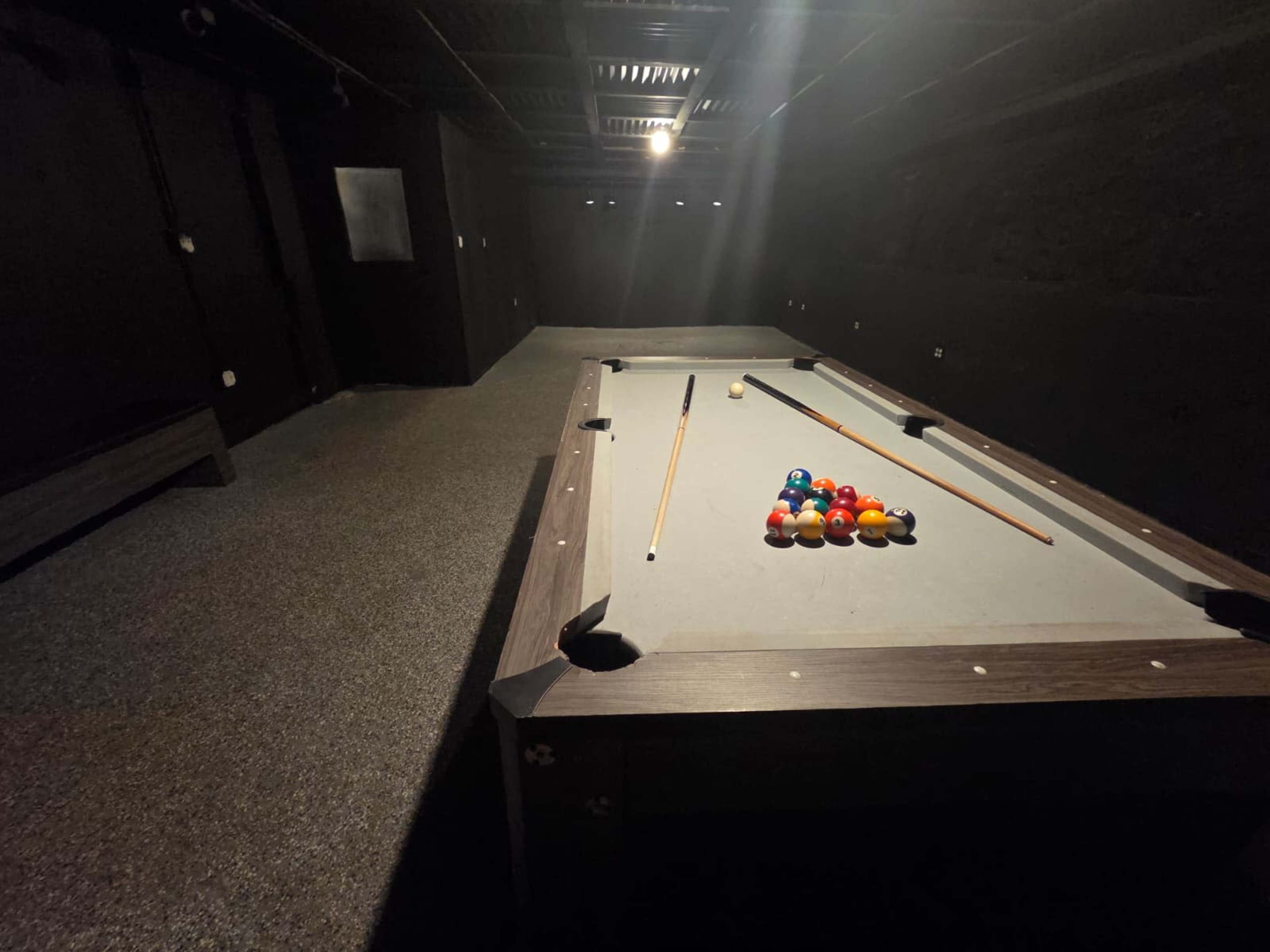The image shows a dimly lit pool room with a billiard table, cue sticks, and a neatly arranged set of colored balls.