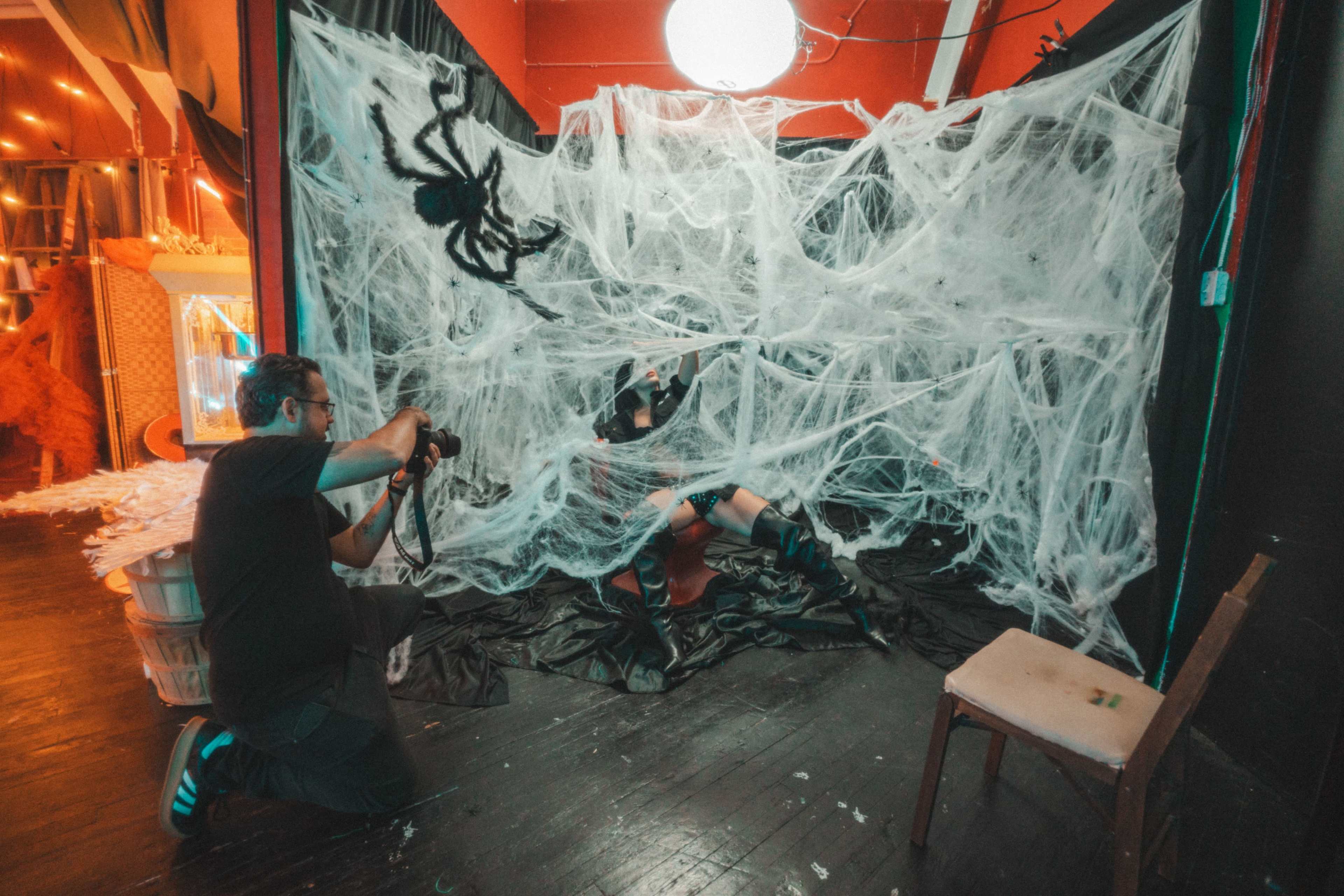 A photographer captures an image of a model posed in front of a spider-web themed backdrop adorned with artificial spider webs and a large spider decoration.