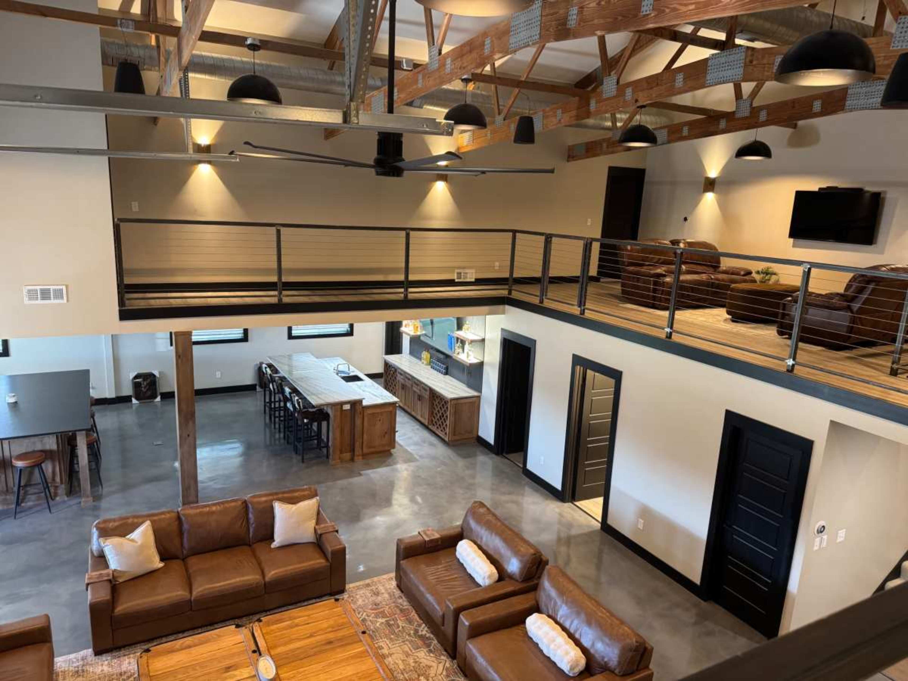 The image shows a spacious interior with a high ceiling, featuring a living area with brown leather sofas, a dining space, and a modern kitchen with an island.