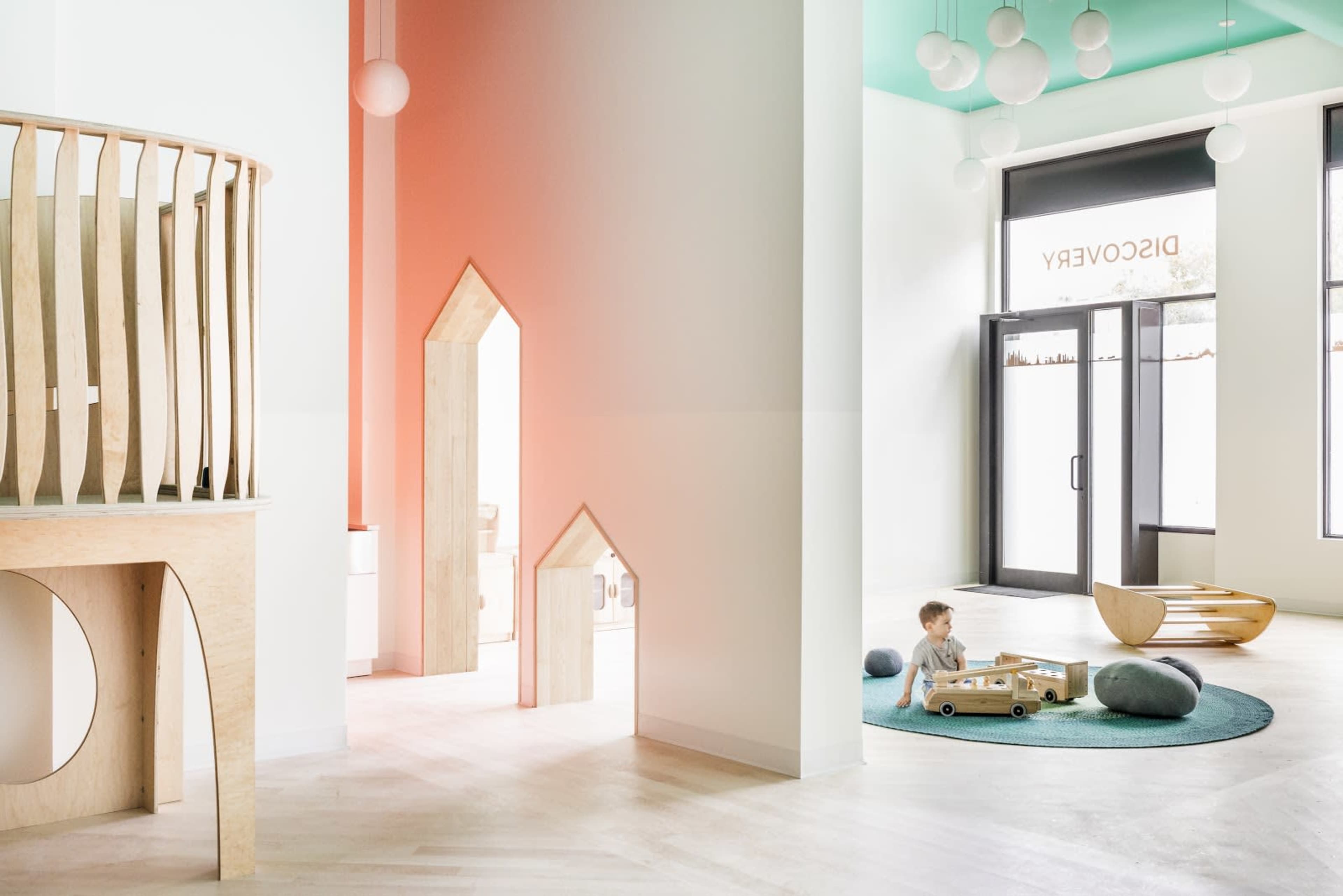 A child plays with a wooden toy car on a circular rug in a brightly lit room featuring wooden architectural elements and playful decor.