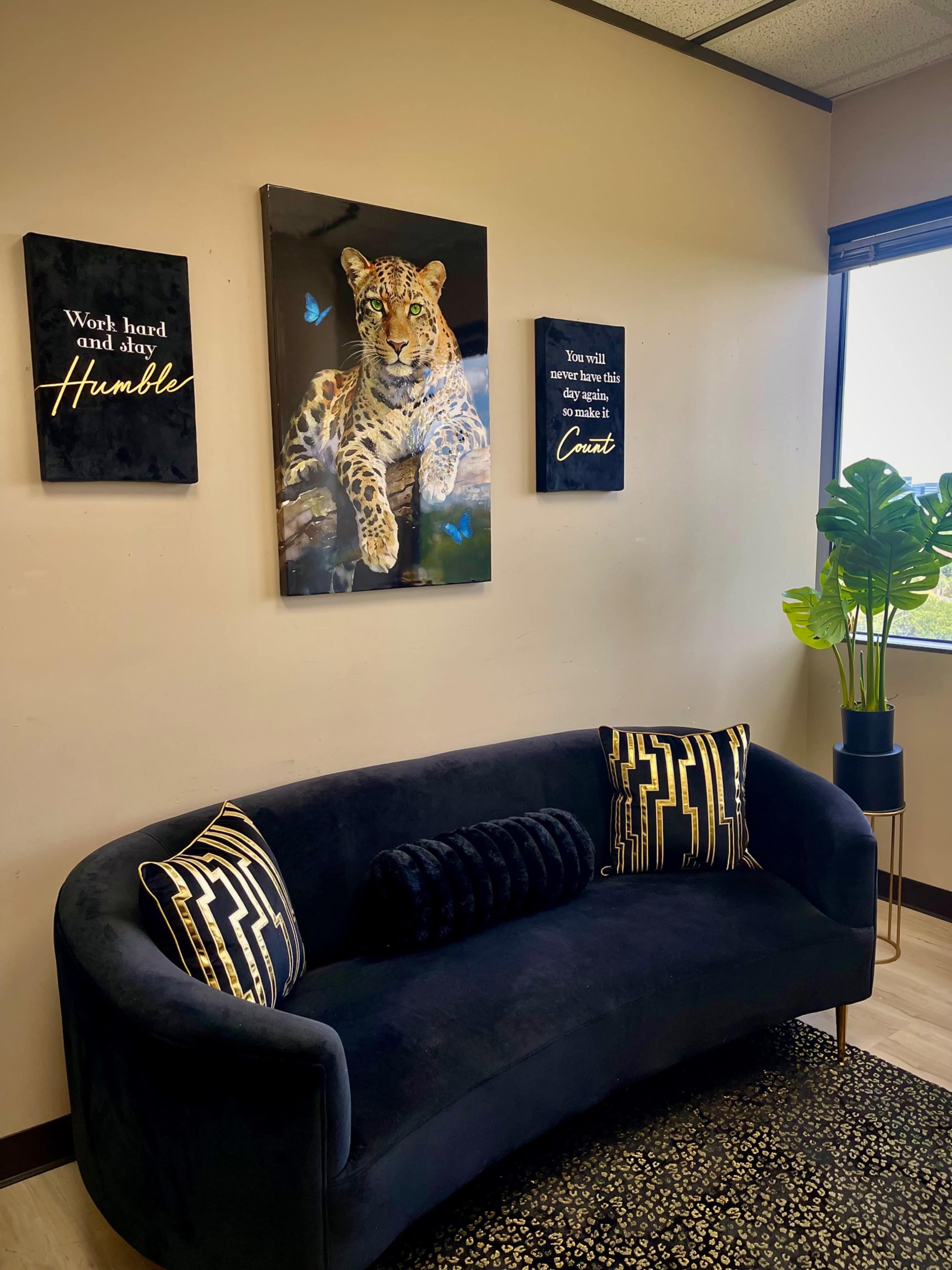 The image shows a cozy office space featuring a black curved sofa with patterned pillows, framed inspirational wall art, and a large portrait of a leopard surrounded by butterflies.