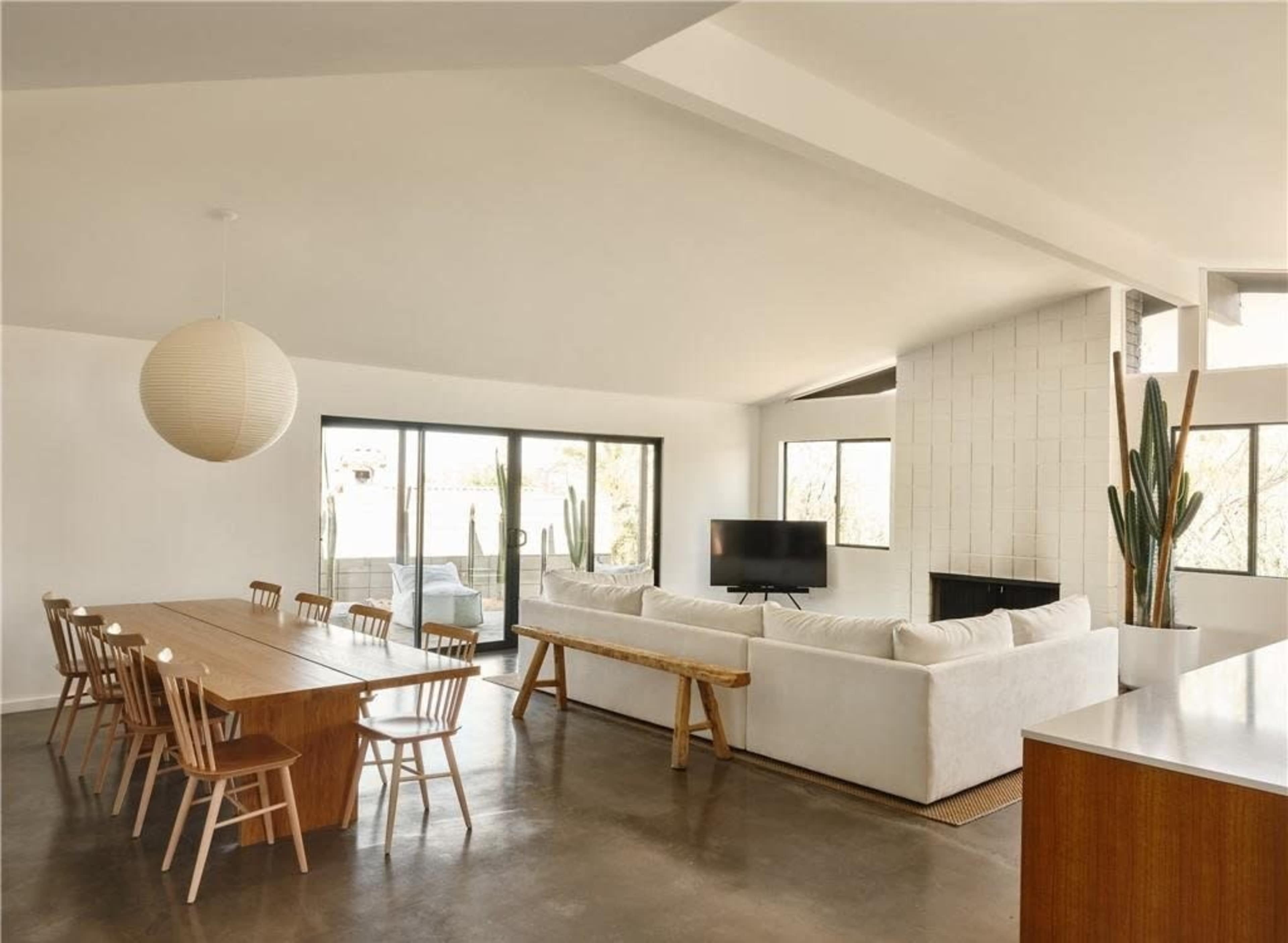A modern living space featuring a dining table with chairs, a sectional sofa, a television, and large windows with views of greenery.
