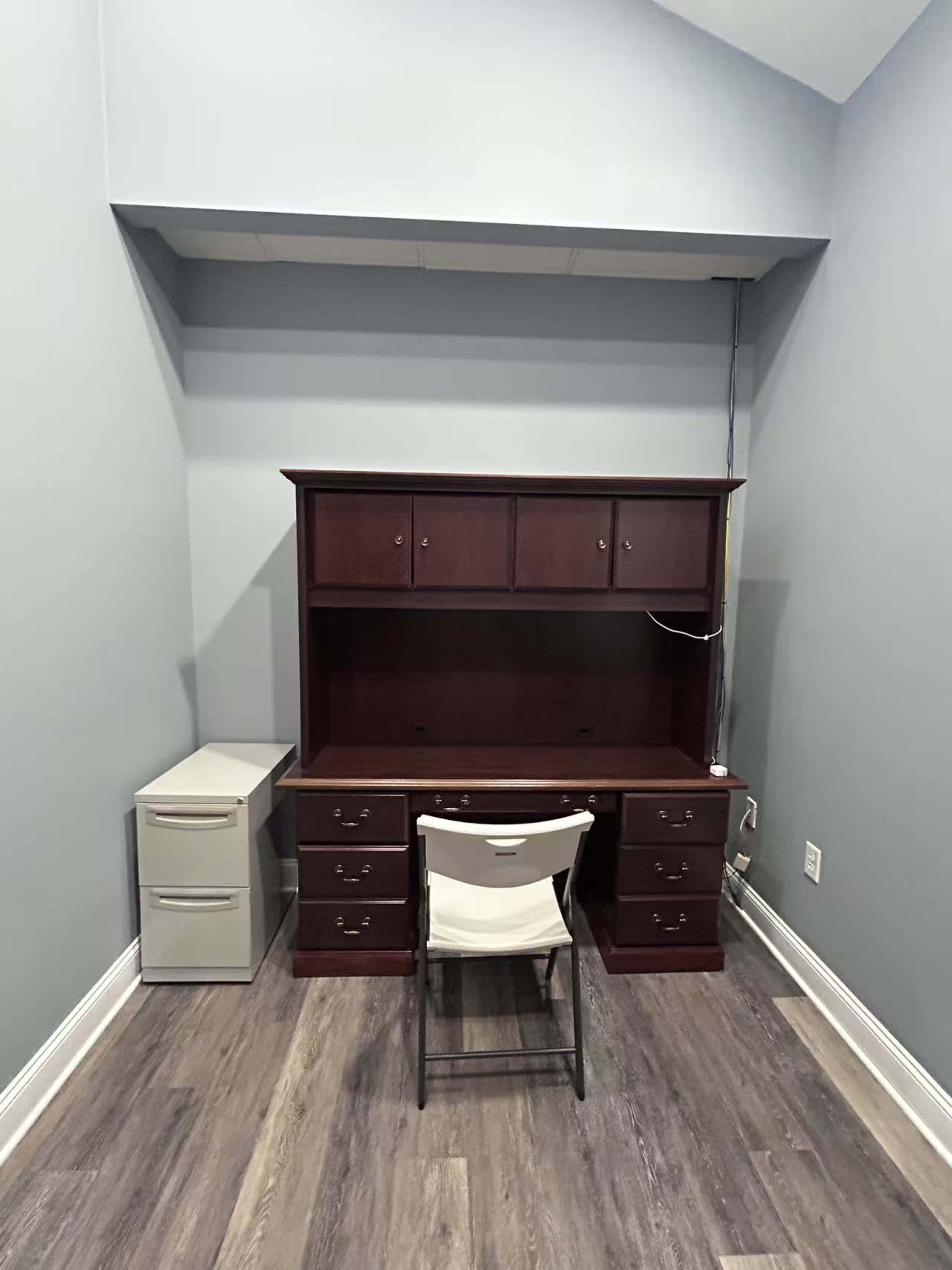A wooden desk with a hutch is positioned against a gray wall, accompanied by a chair and a small filing cabinet nearby.