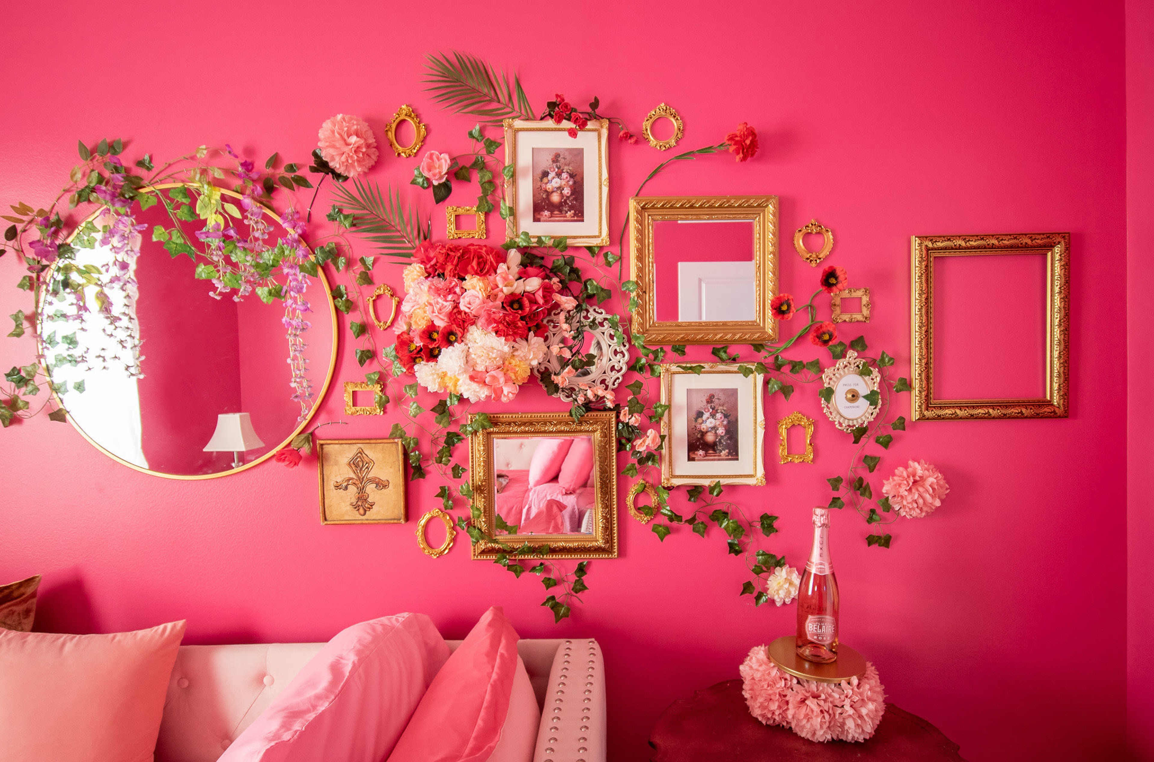 The scene features a vibrant pink wall adorned with floral decorations, framed photos, and a round mirror, alongside a sofa with pink pillows and a tabletop embellished with flowers and a bottle.