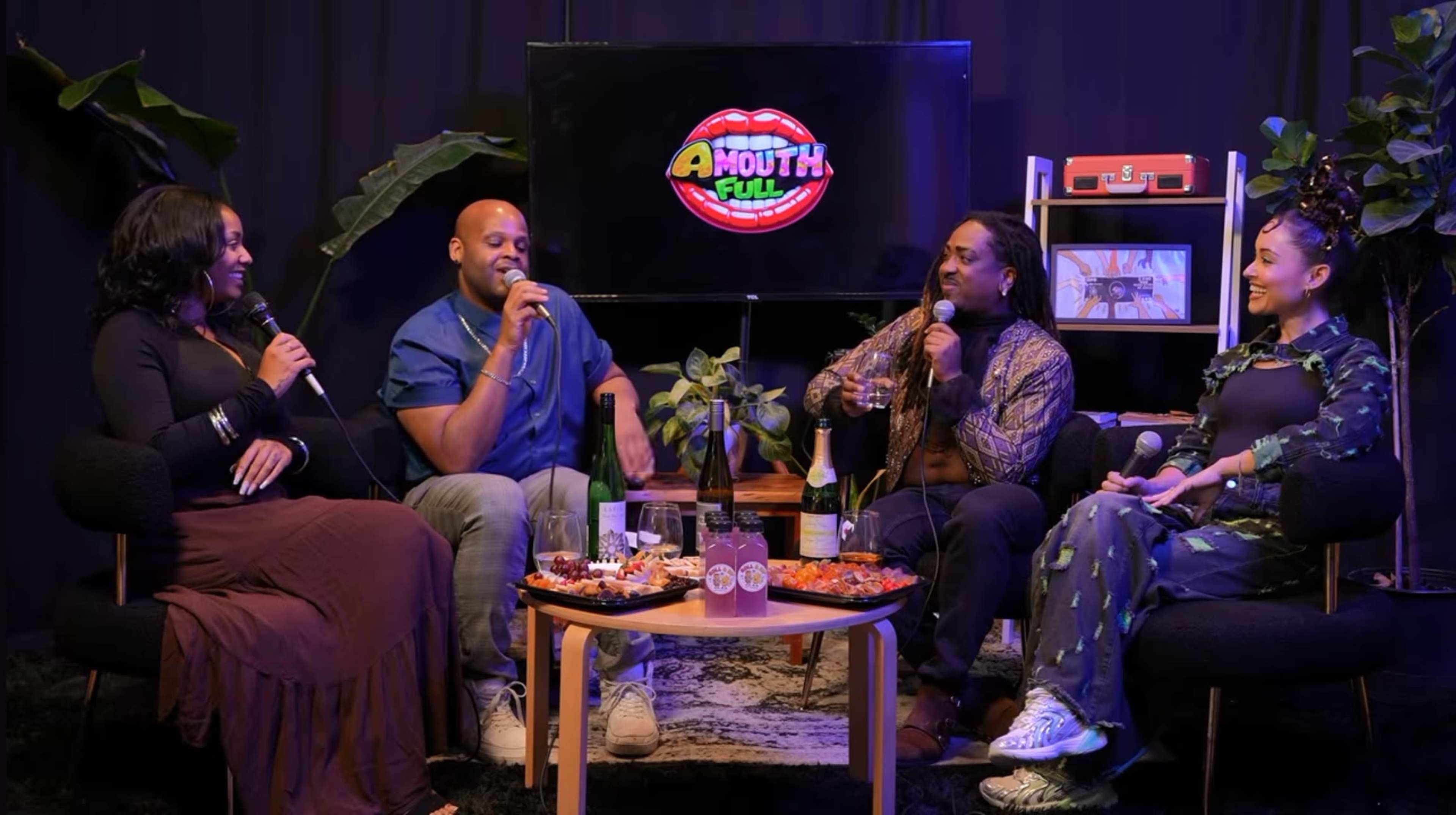 A group of four people sits in a room, engaging in conversation while seated around a coffee table filled with food and drinks, with a television in the background displaying a colorful logo.