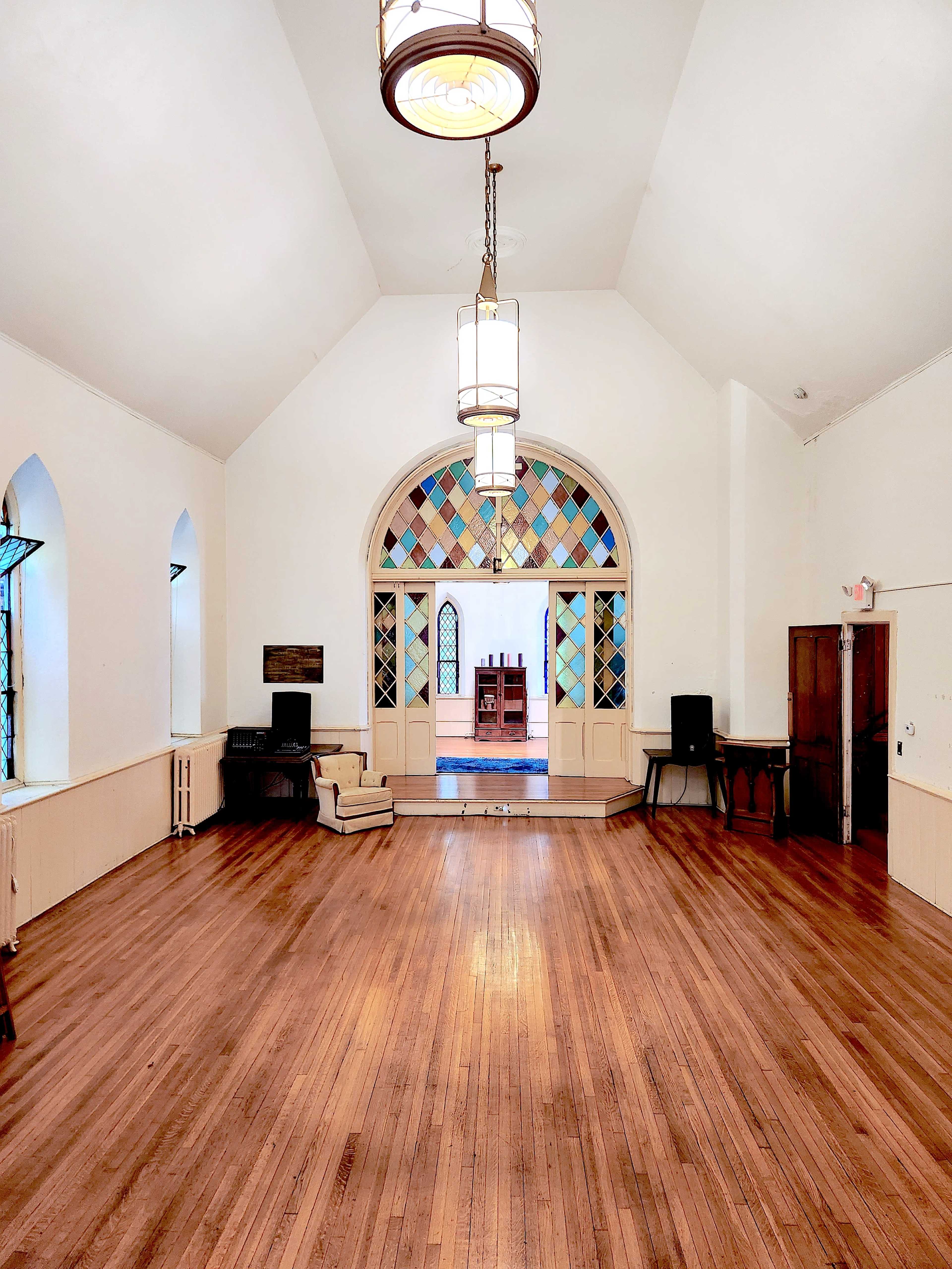 The interior of a spacious, well-lit room features hardwood floors and large arched windows with colorful panes.